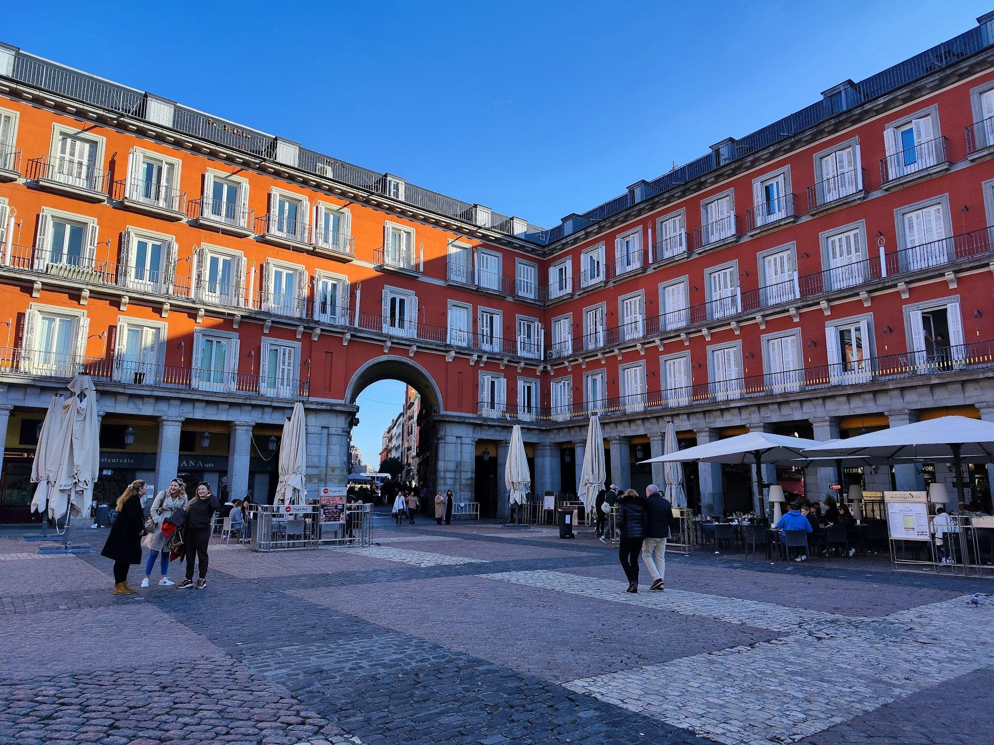 People walking across Plaza Mayor in Madrid, with red facades, an arched gateway and café umbrellas under the arcades.