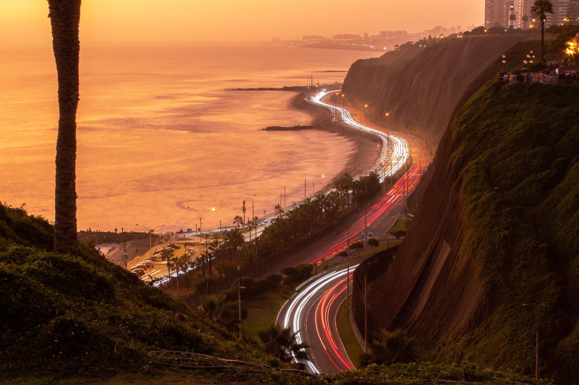 The Costa Verde road curving below the Miraflores cliffs in Lima at sunset, with light trails from cars.