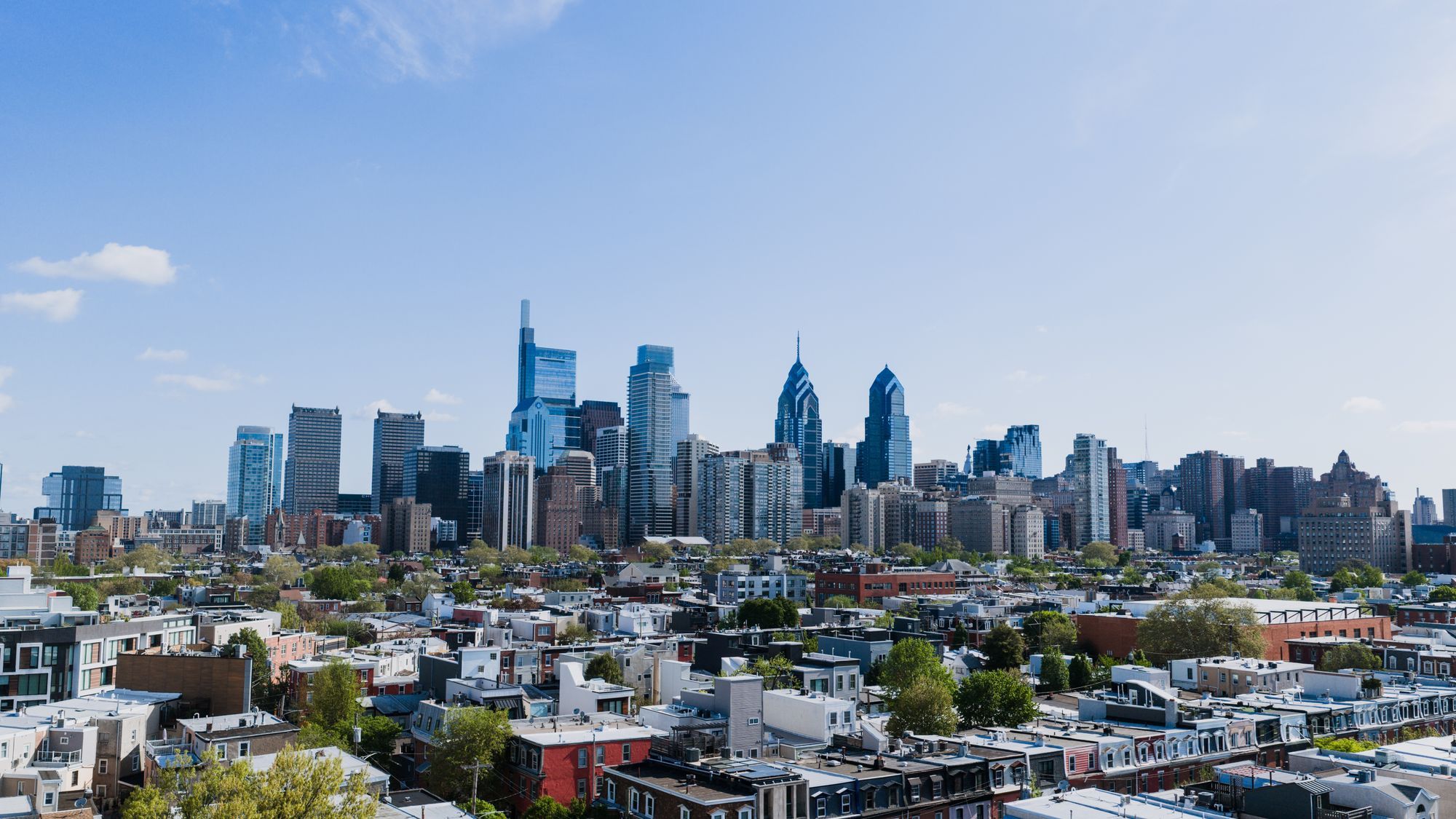 The Center City skyline of Philadelphia rising over rows of rooftops under a blue sky.