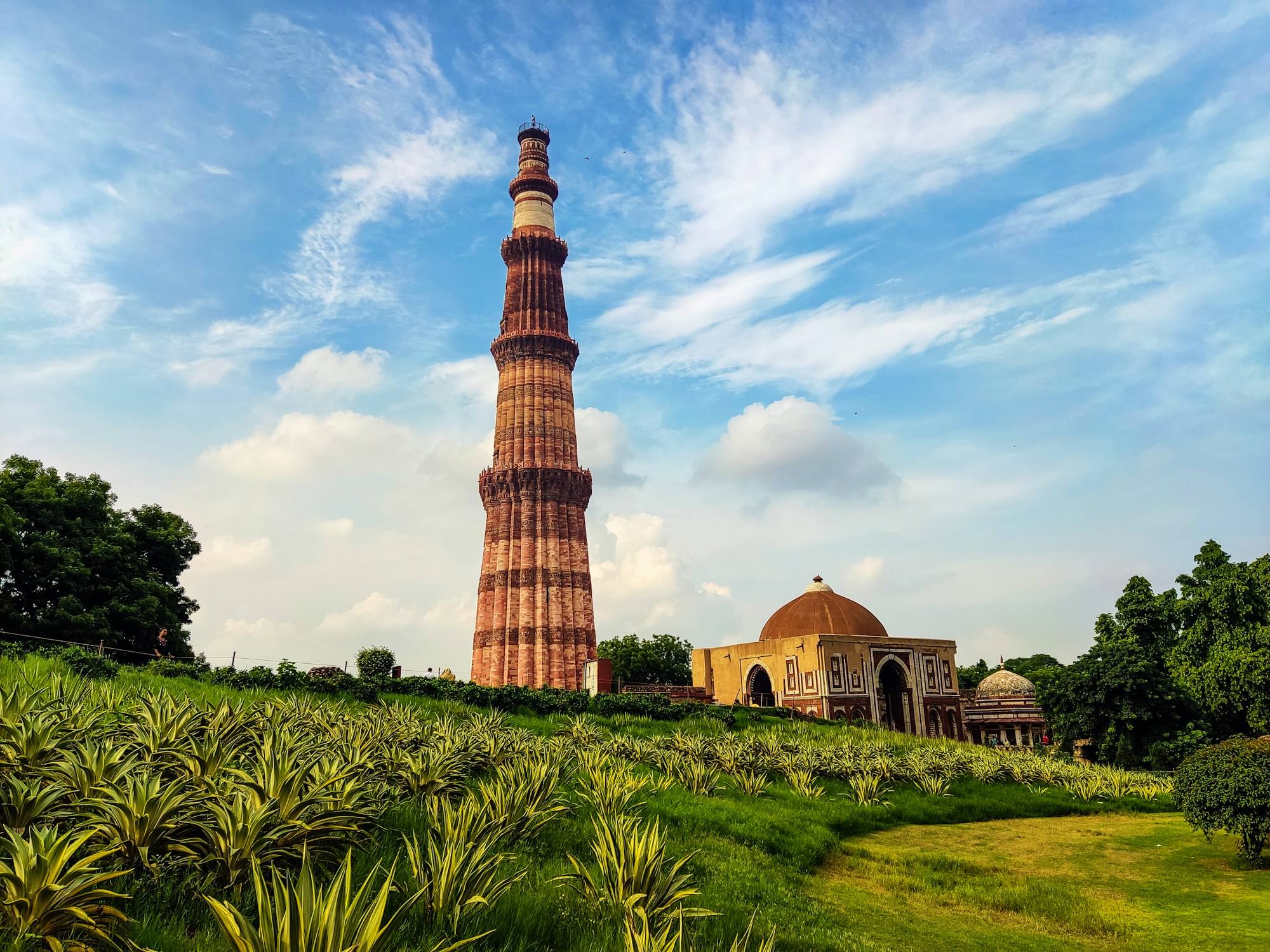 The Qutub Minar rising above a green lawn in Mehrauli, New Delhi, under a blue sky streaked with cloud.