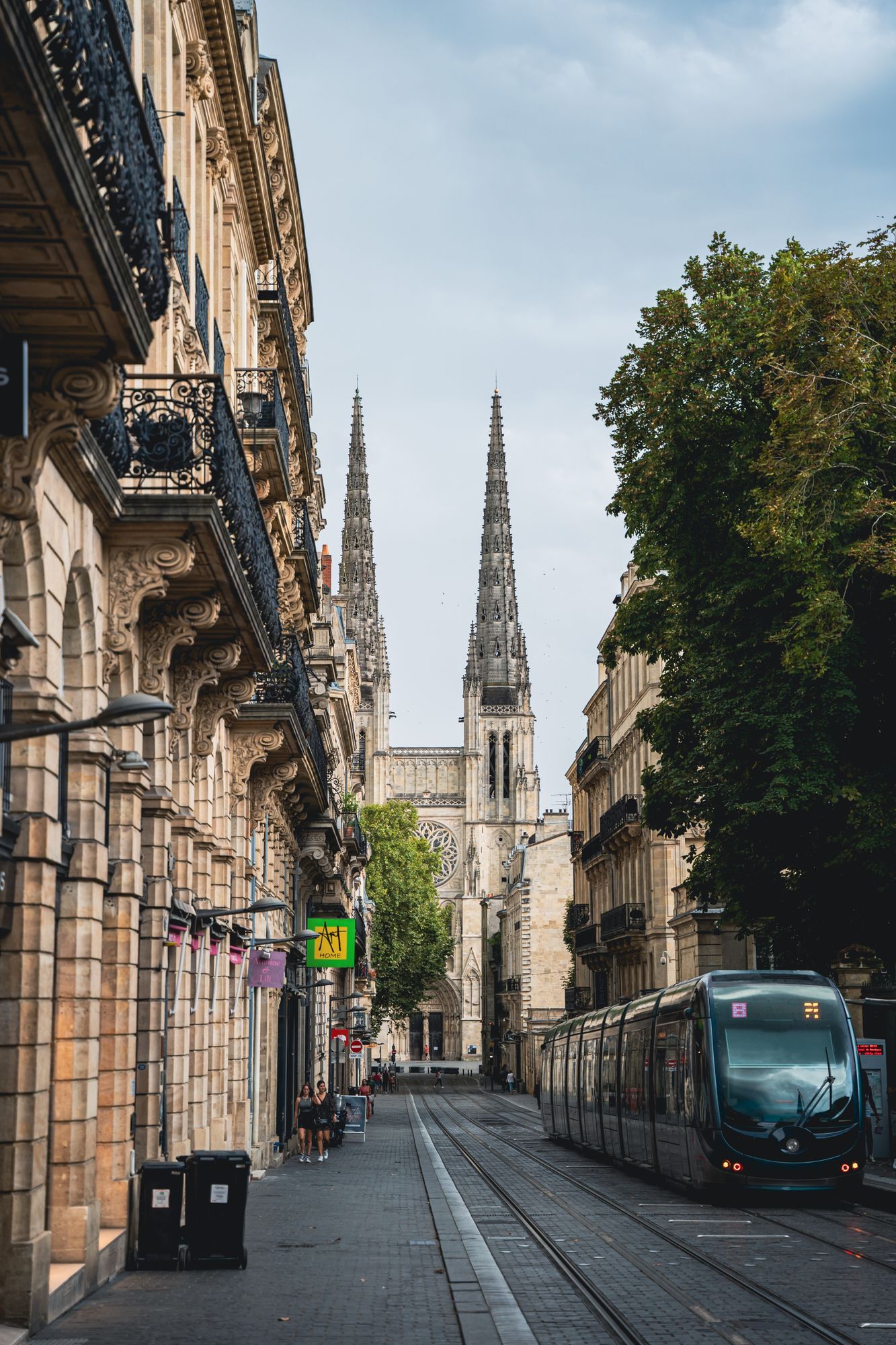 A street of balconied stone buildings in Bordeaux with a tram and the spires of Saint-André cathedral at the end.