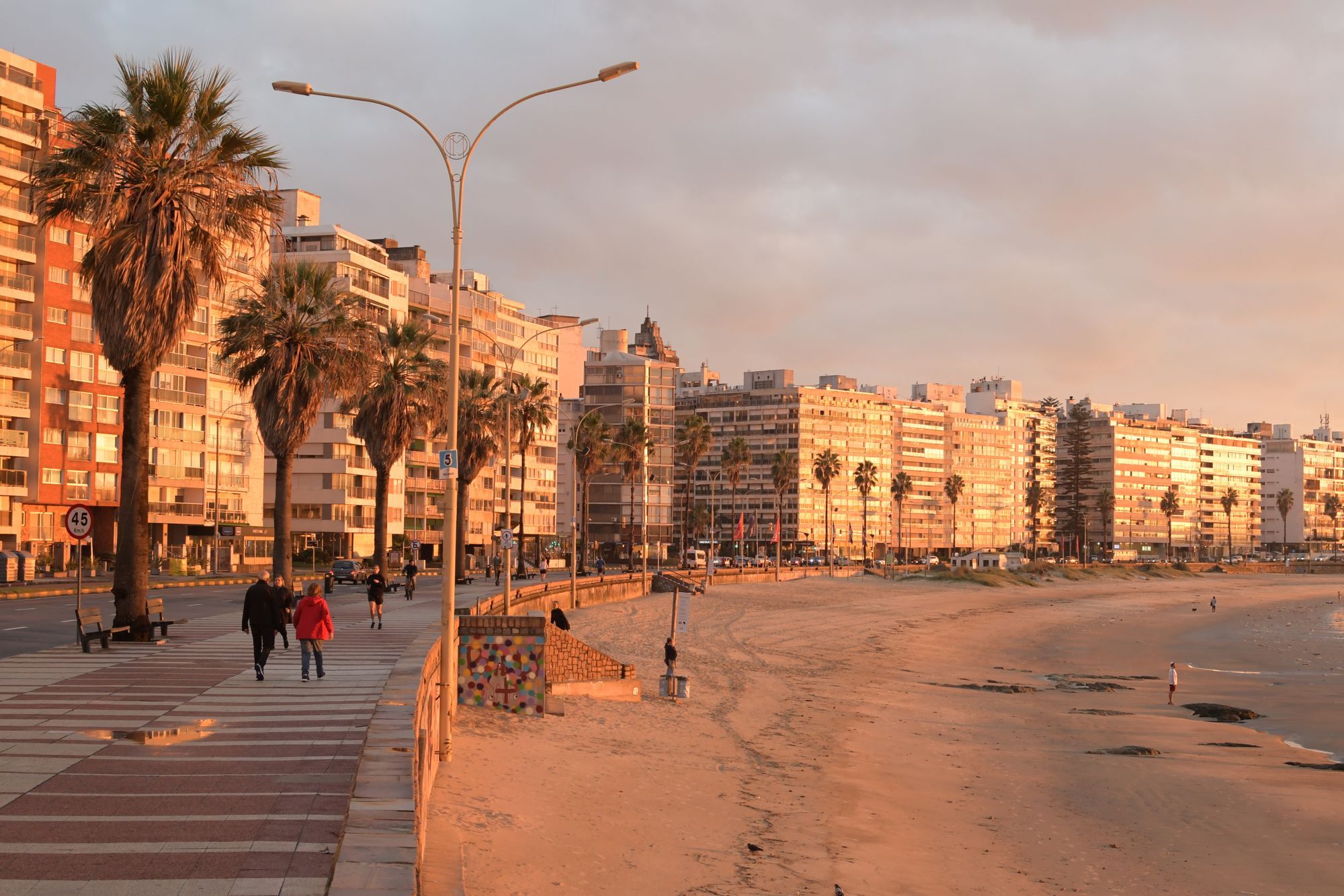 People walking along the Rambla beside the beach in Montevideo, with palm trees and apartment blocks in warm evening light.