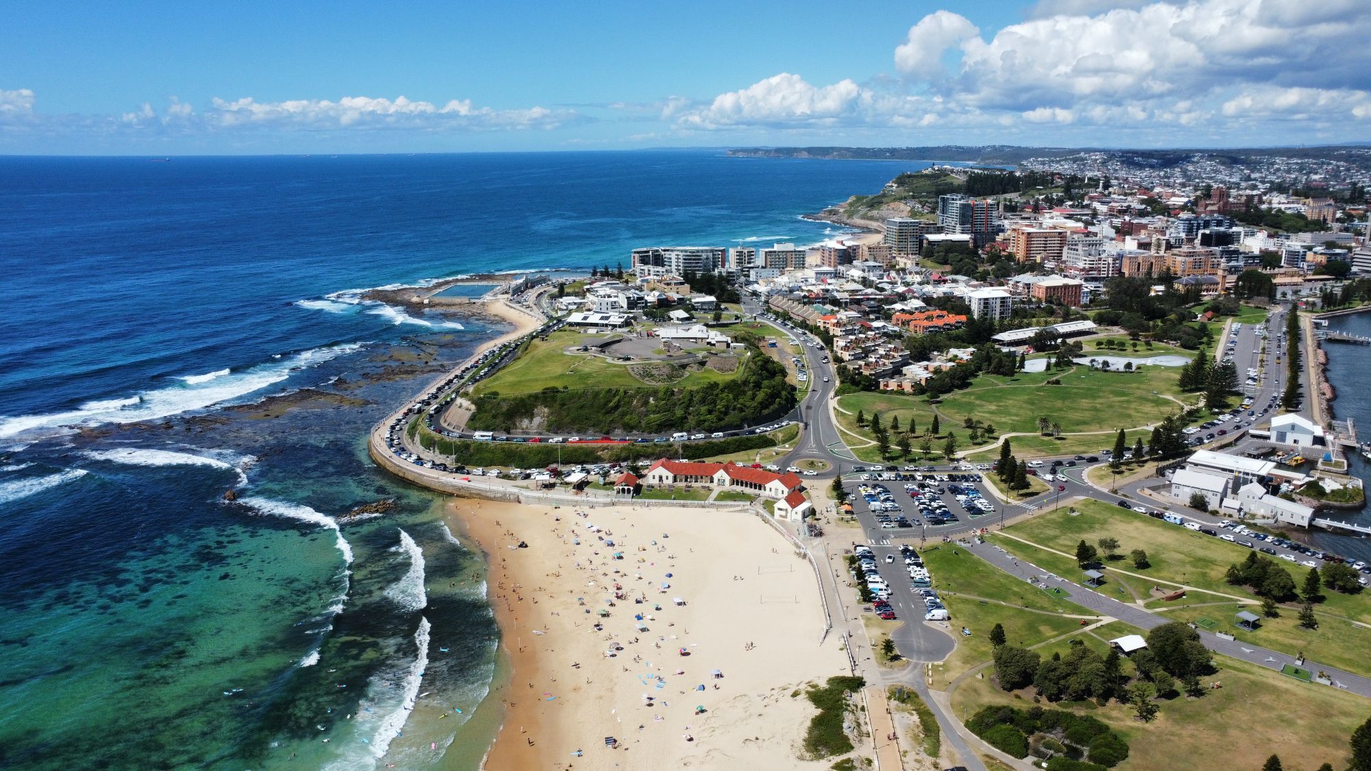 An aerial view of Newcastle Beach and the east end of the city, with surf on the left and the town behind a green headland.