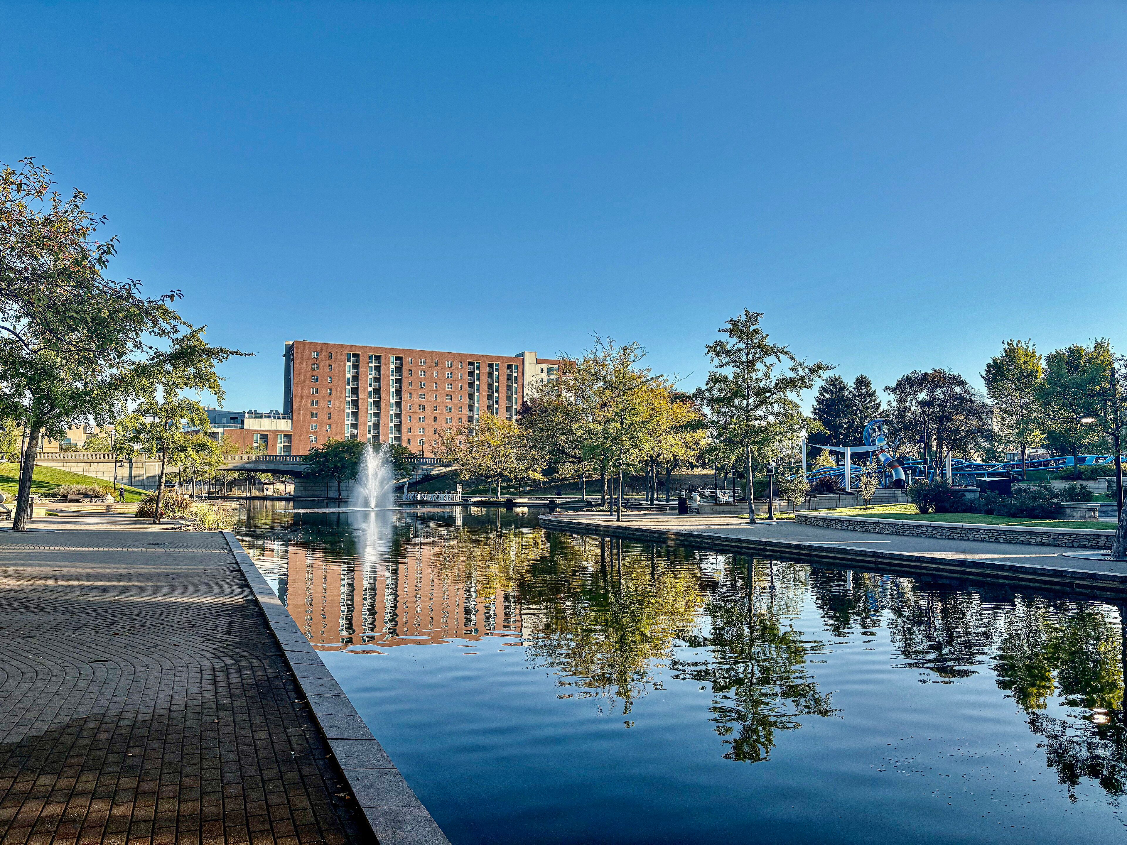 The Canal Walk in downtown Indianapolis, with a fountain and trees reflected in the water.