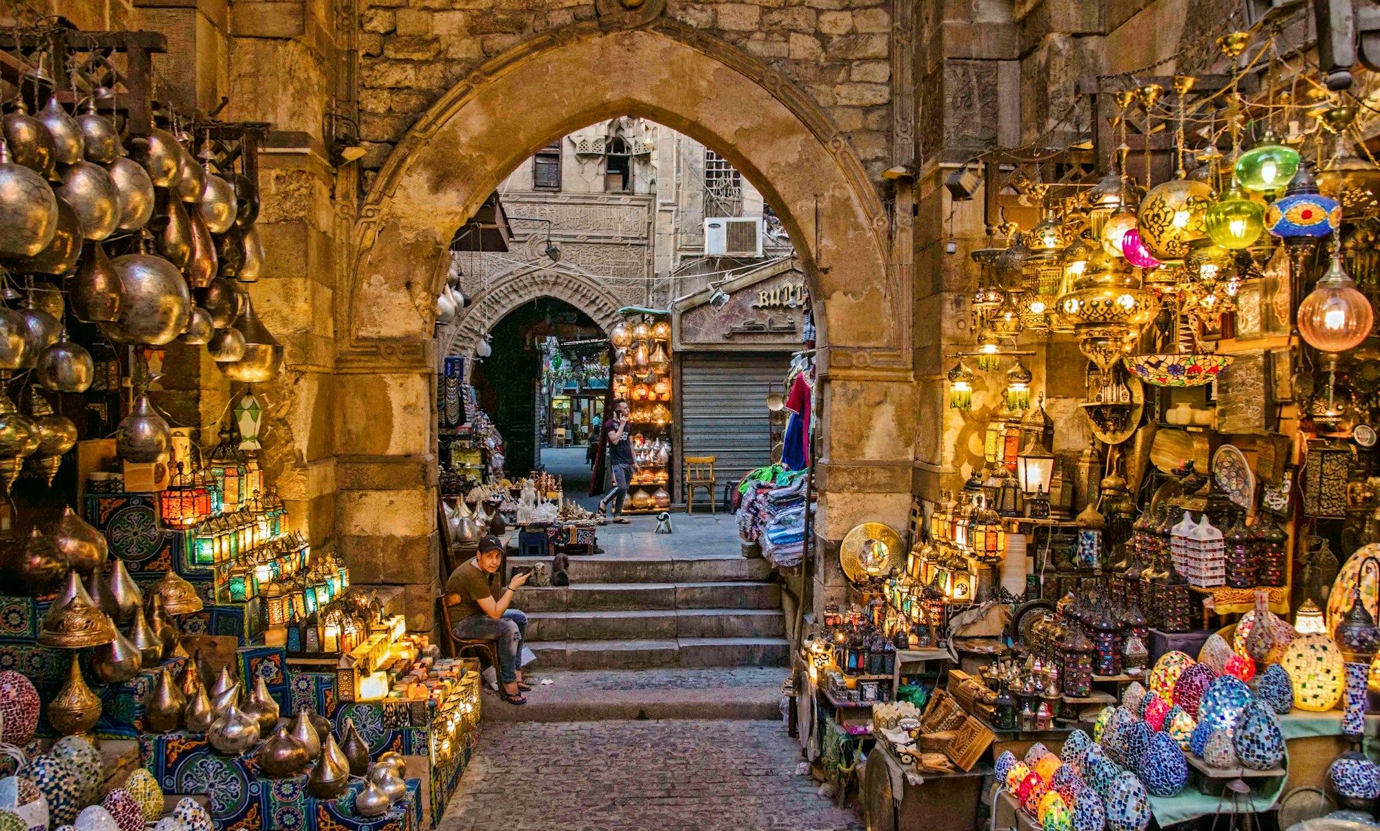 A stone archway in the Khan el-Khalili bazaar in Cairo, with brass lanterns and glass lamps on sale along both sides of the alley.