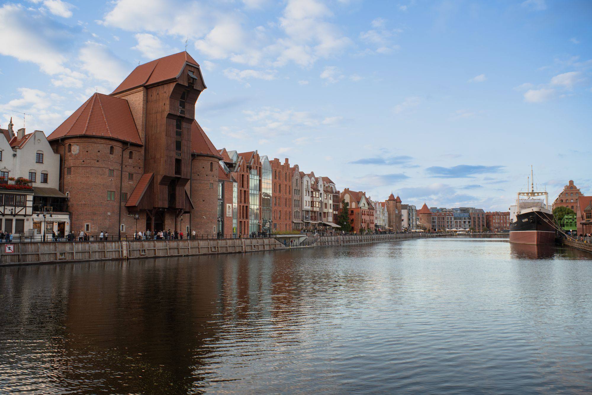 The medieval Crane on the Motława river in Gdańsk, with the old town buildings along the quay and calm water in the foreground.