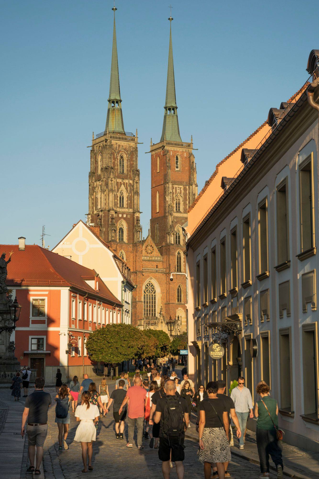 The twin towers of Wrocław Cathedral on Ostrów Tumski above a street of walkers in warm evening light.