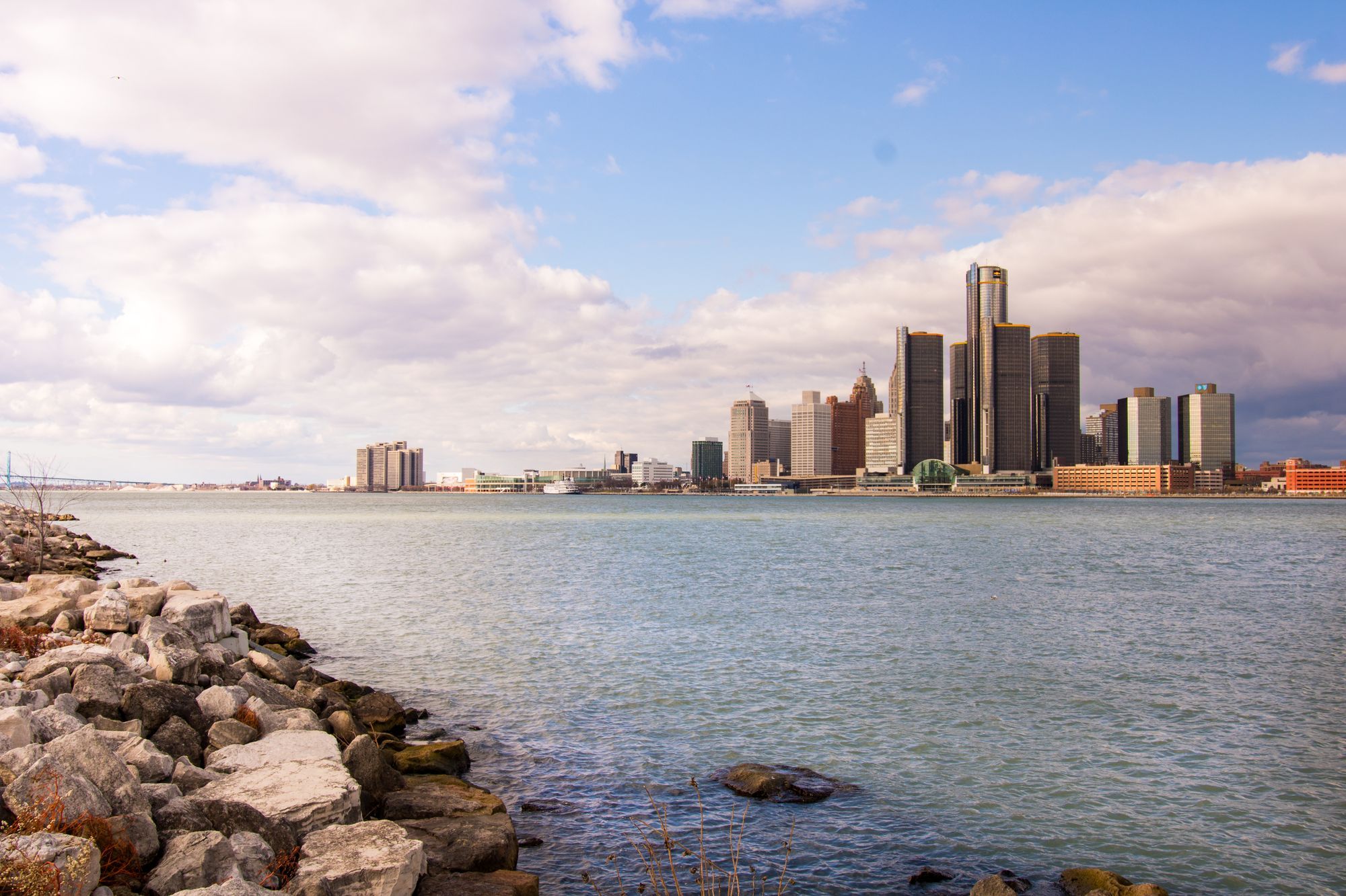 The Detroit skyline across the river, seen over a rocky shore.