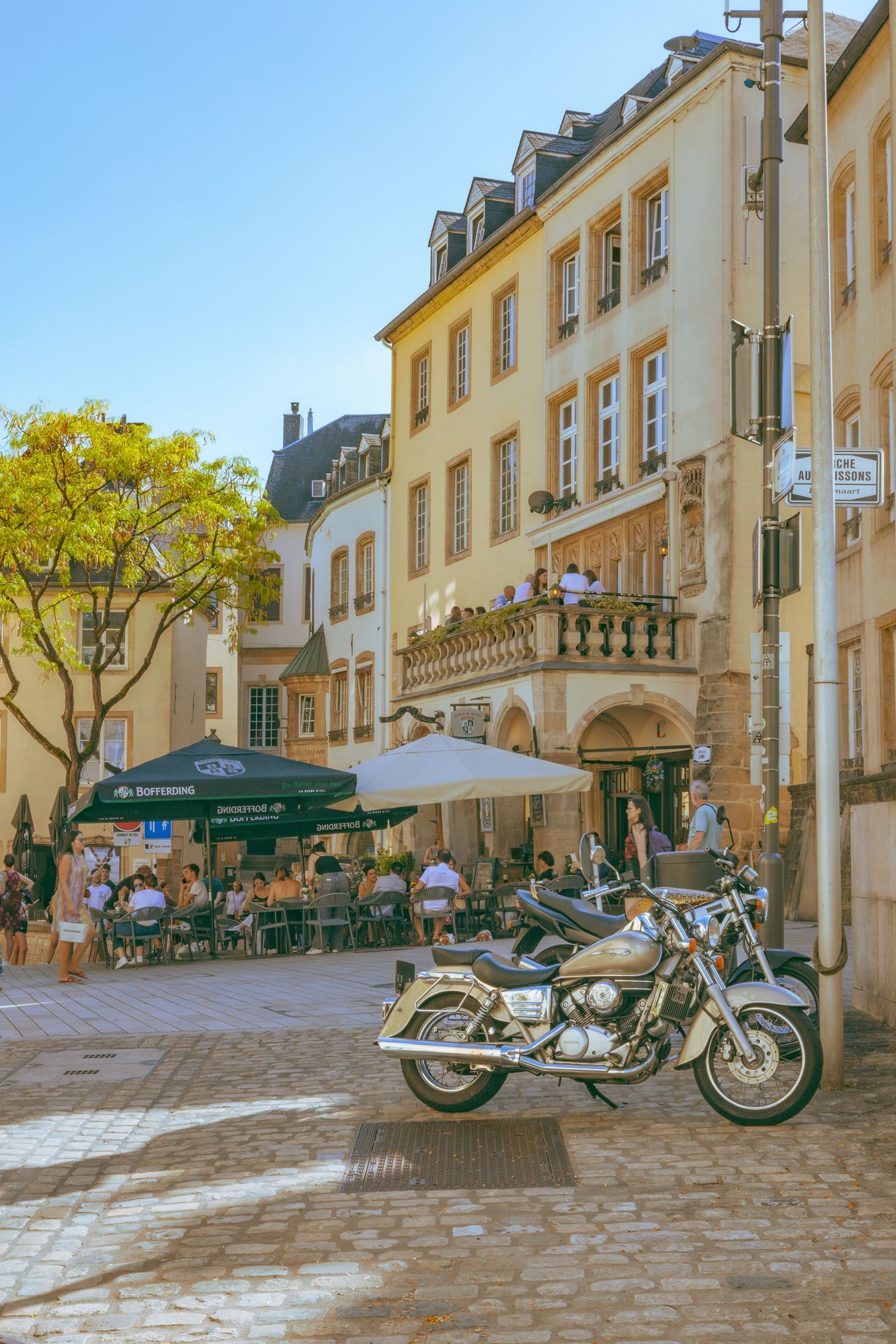 A café terrace with people at tables and a parked motorcycle on a cobbled street in Luxembourg City.