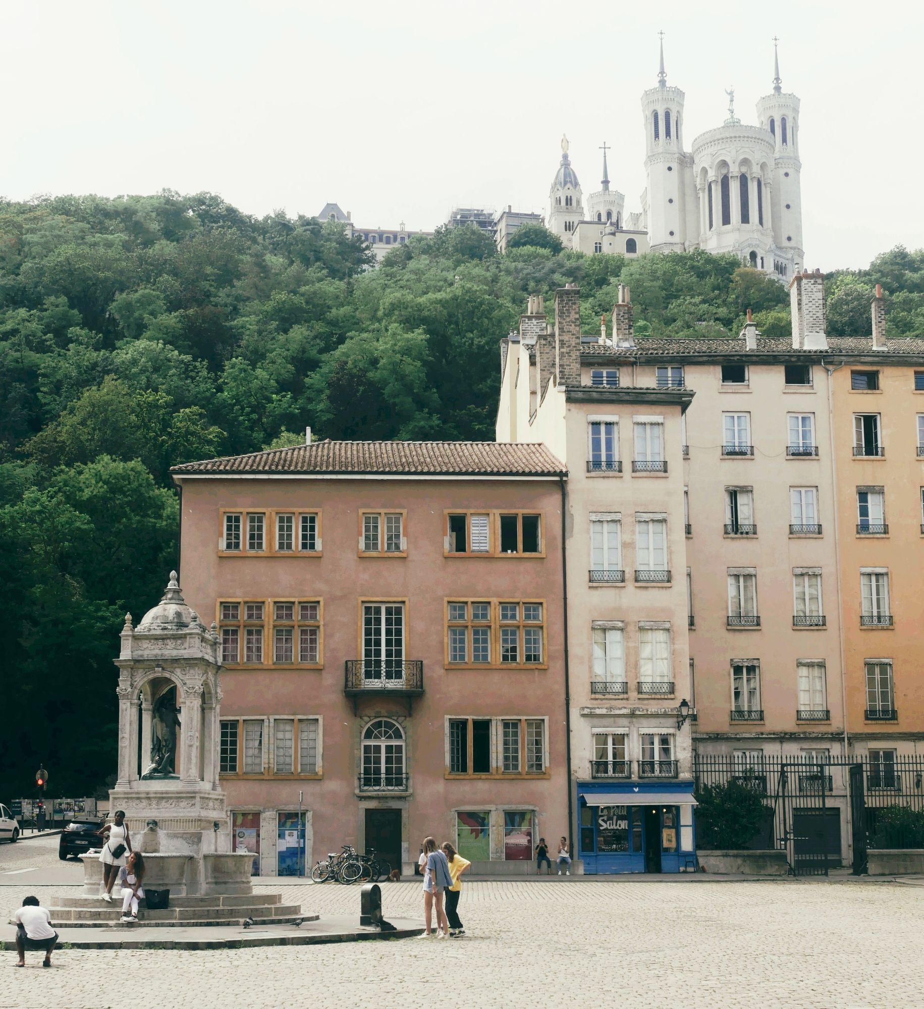A small square in old Lyon with pale and pink facades, a stone fountain and the Basilica of Notre-Dame de Fourvière above the trees.