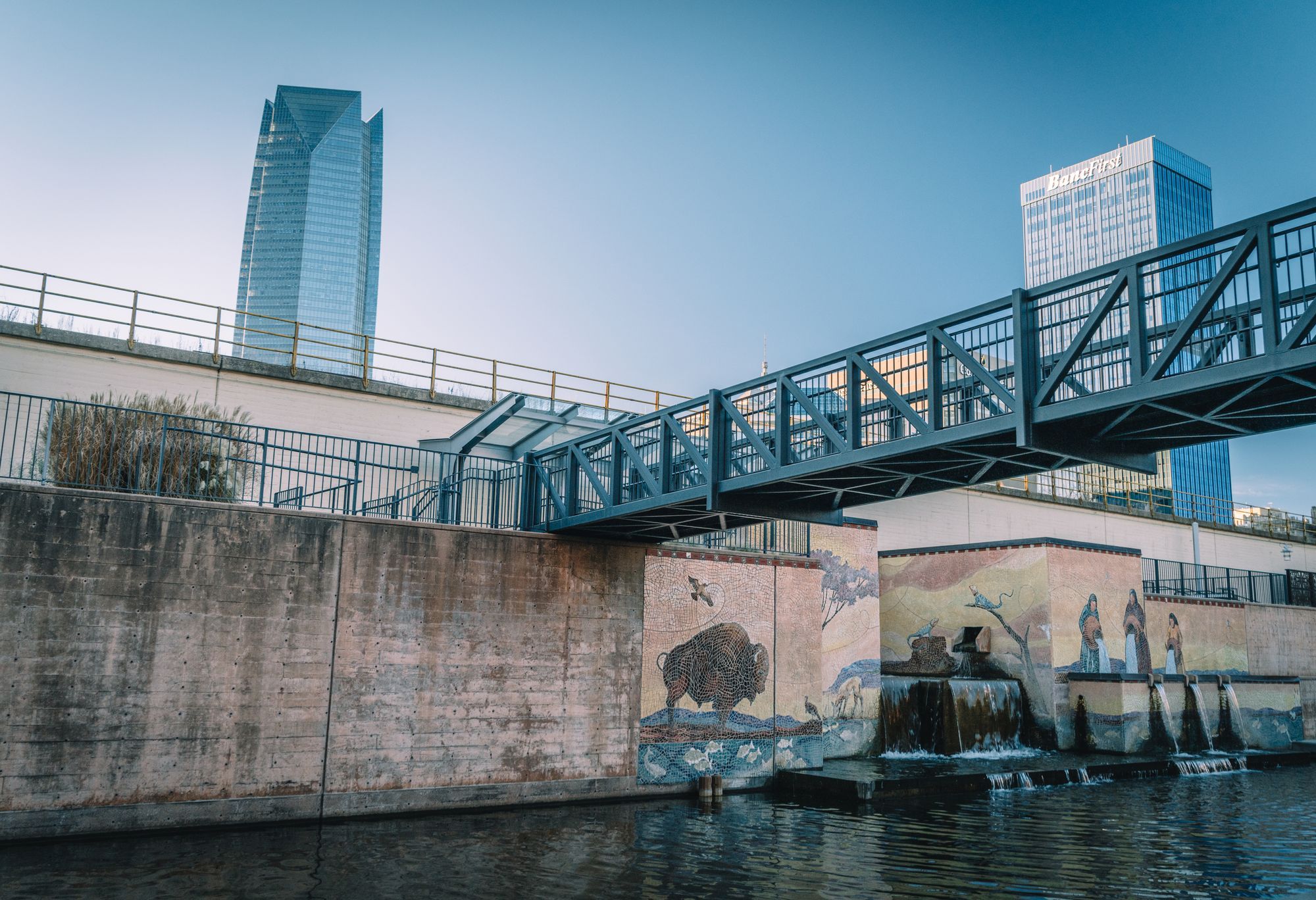 A steel footbridge and mural beside the Bricktown canal in Oklahoma City, with towers behind.