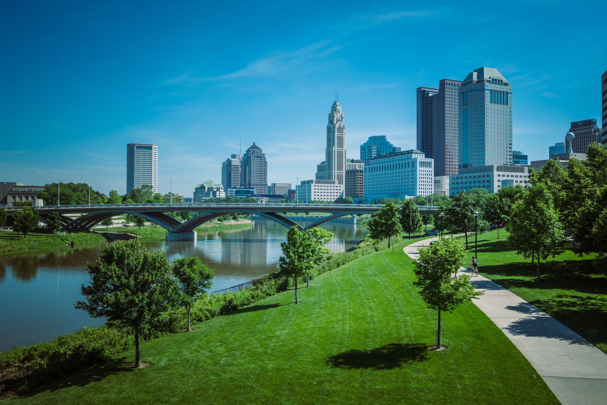 The Columbus skyline over the Scioto River, with a grass riverbank and a path in the foreground.