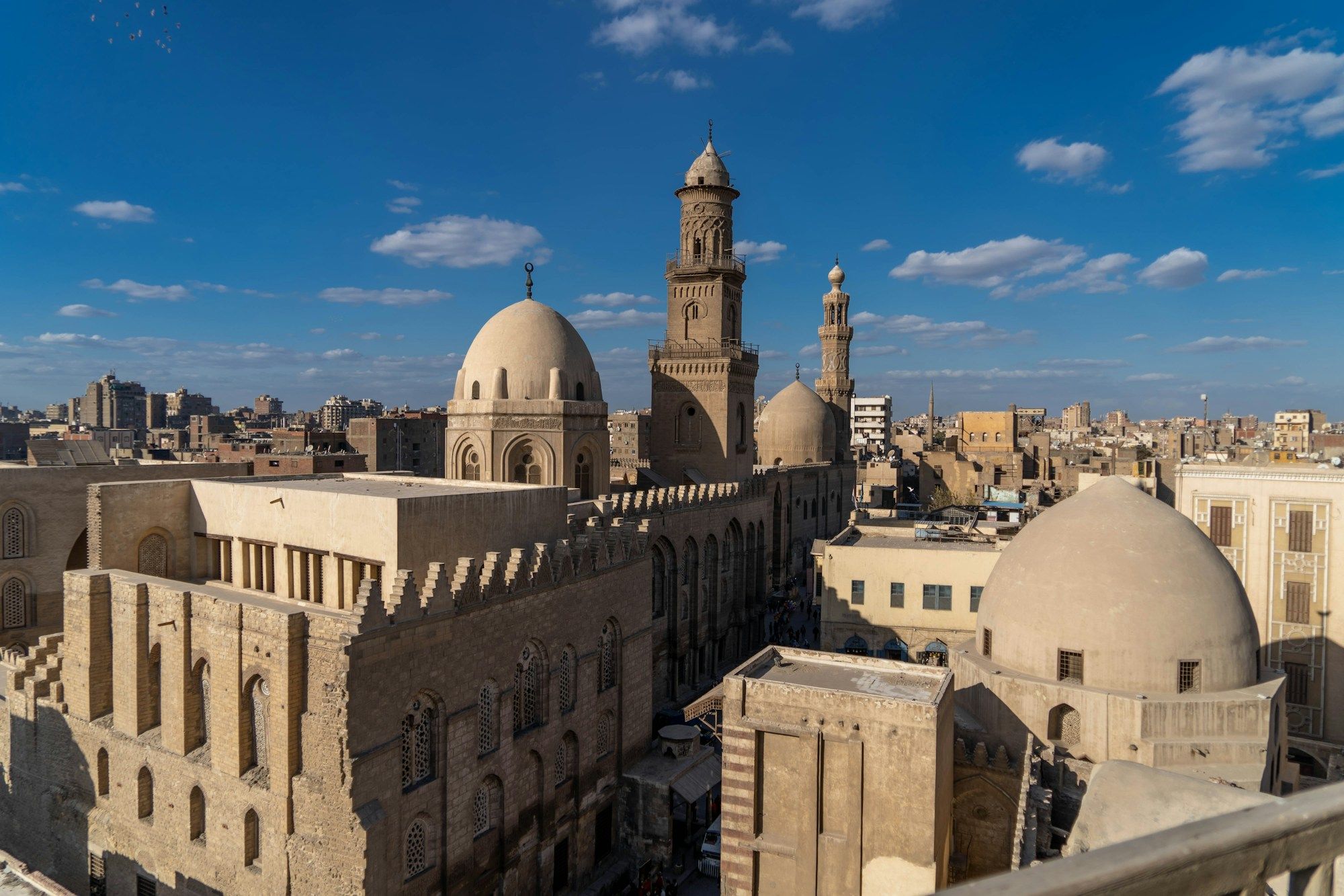 Domes and minarets of Old Cairo seen from a rooftop under a blue sky, with the city stretching out behind them.