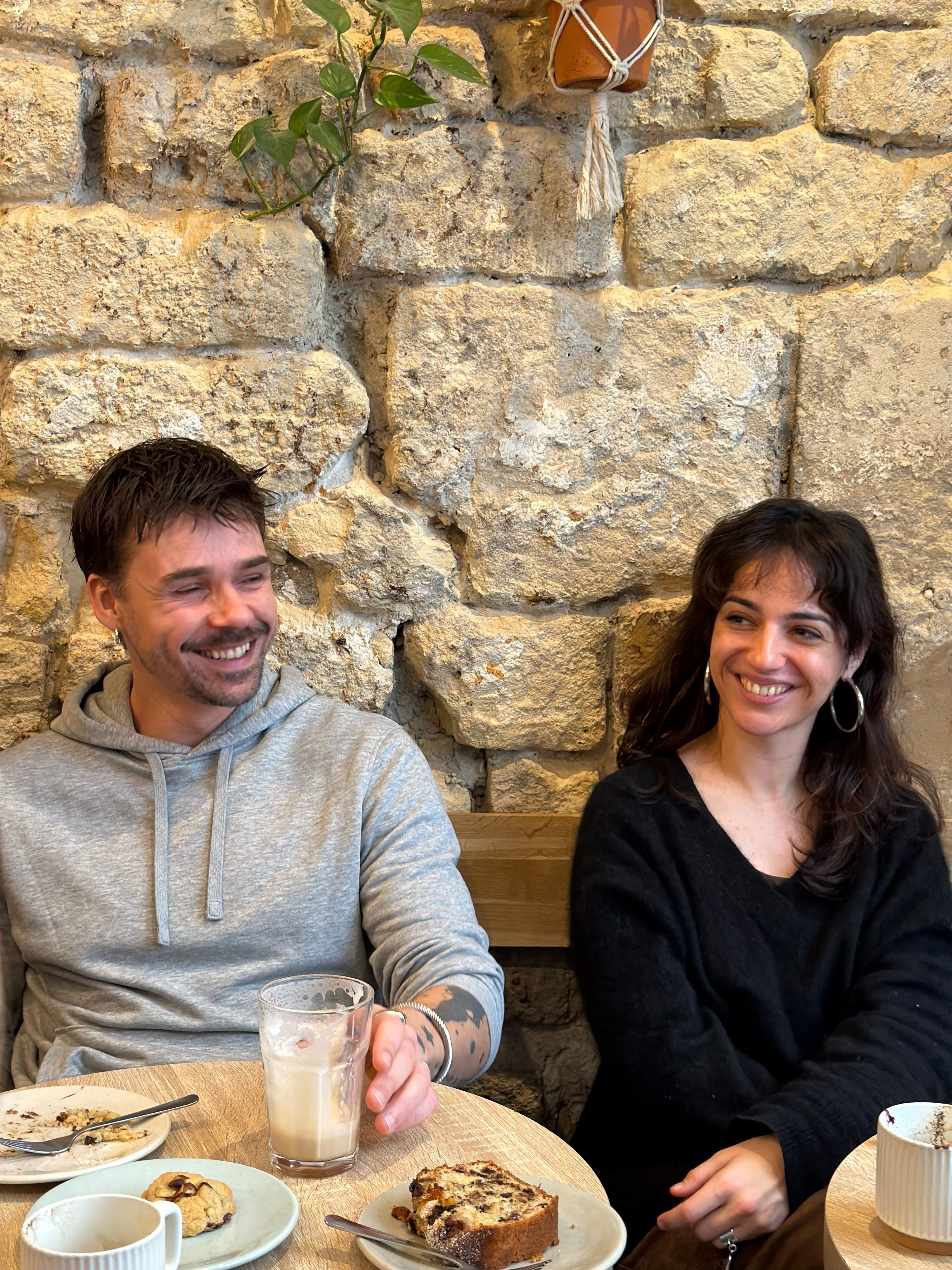 Two people smiling over coffee and cake against a stone café wall