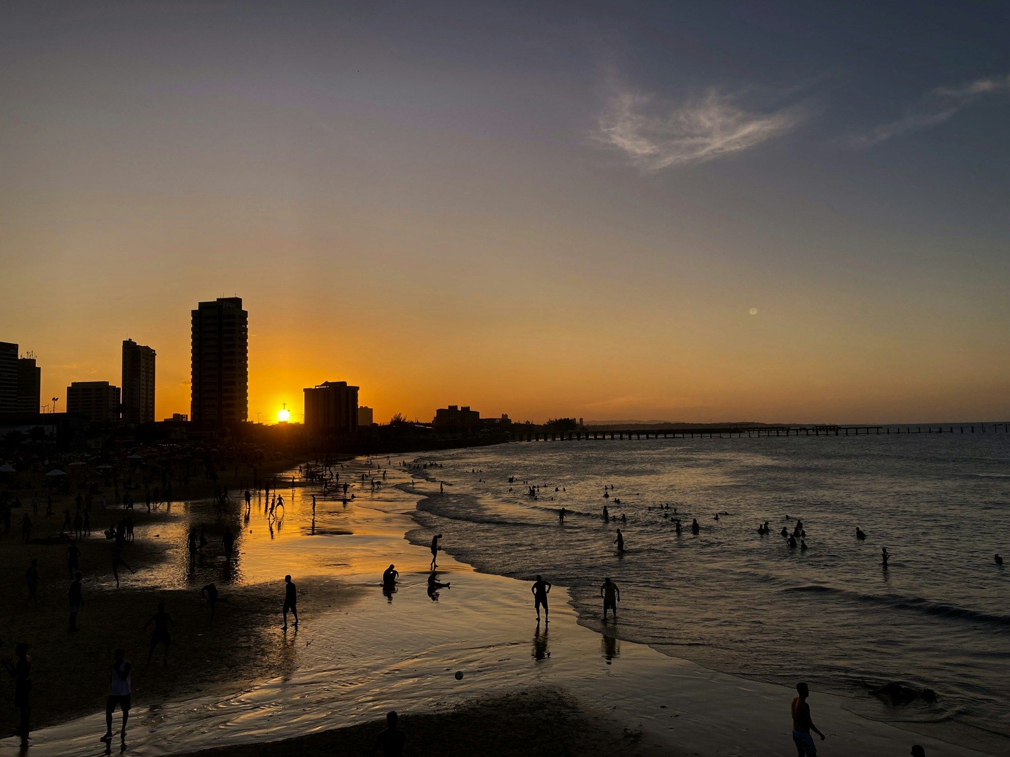 People on the beach in Fortaleza at sunset, with the sun going down behind the city's buildings and a long pier in the distance.