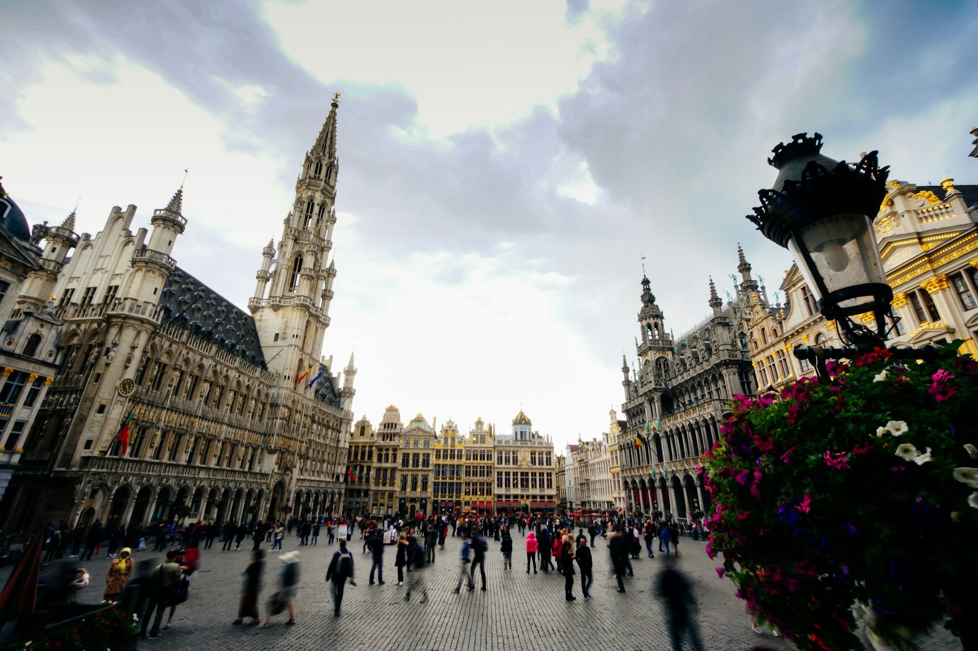 People walking across the Grand Place in Brussels, with the town hall tower behind a street lamp and flowers