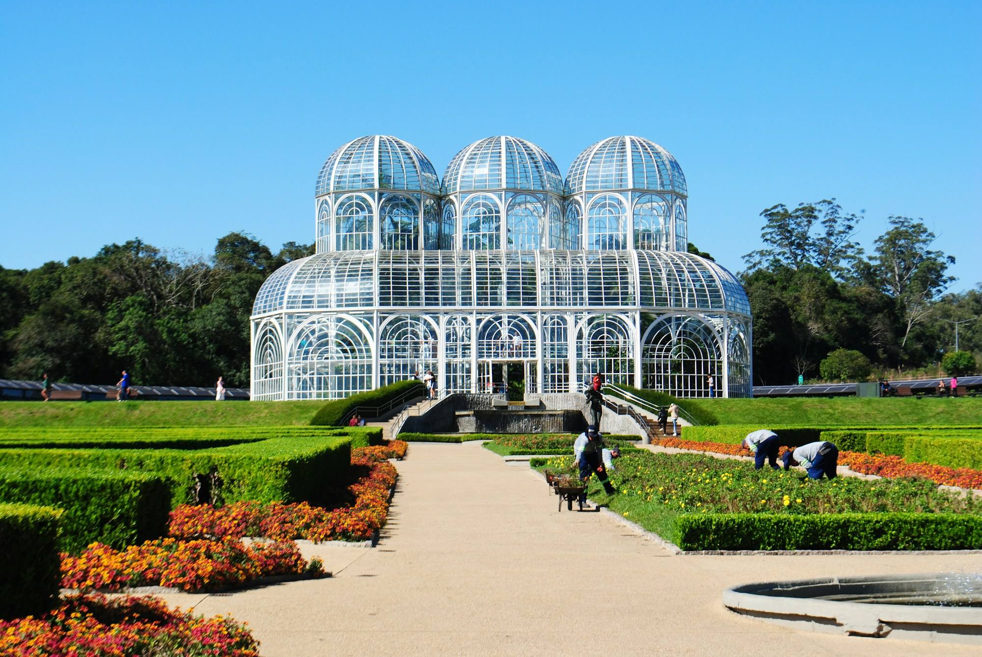 The glass greenhouse of the Jardim Botânico in Curitiba under a blue sky, with clipped hedges and flower beds in front.