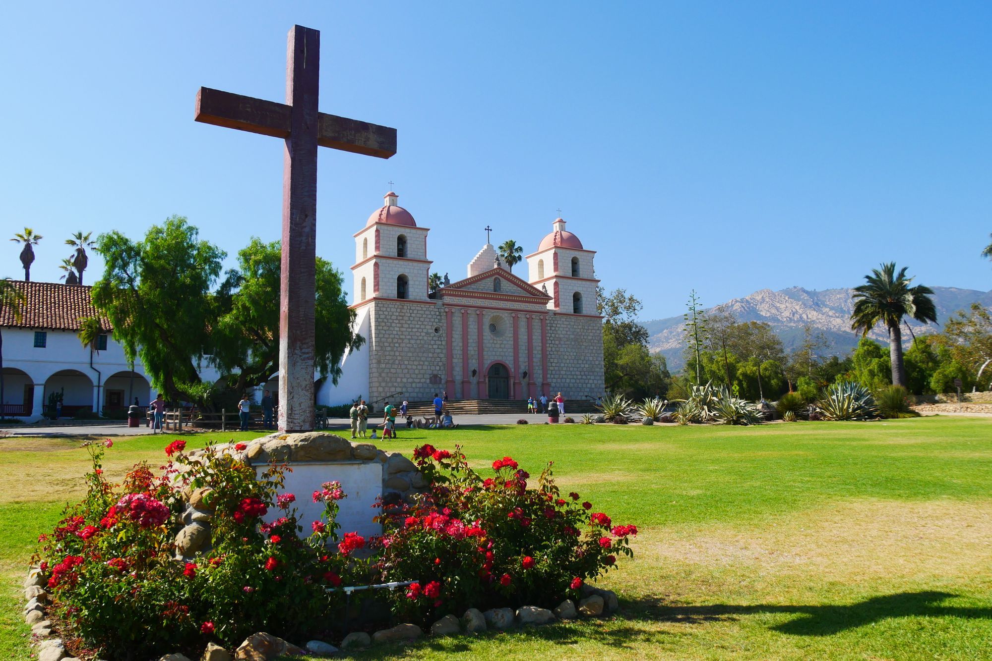 Old Mission Santa Barbara with its two towers behind a wooden cross and red flowers.