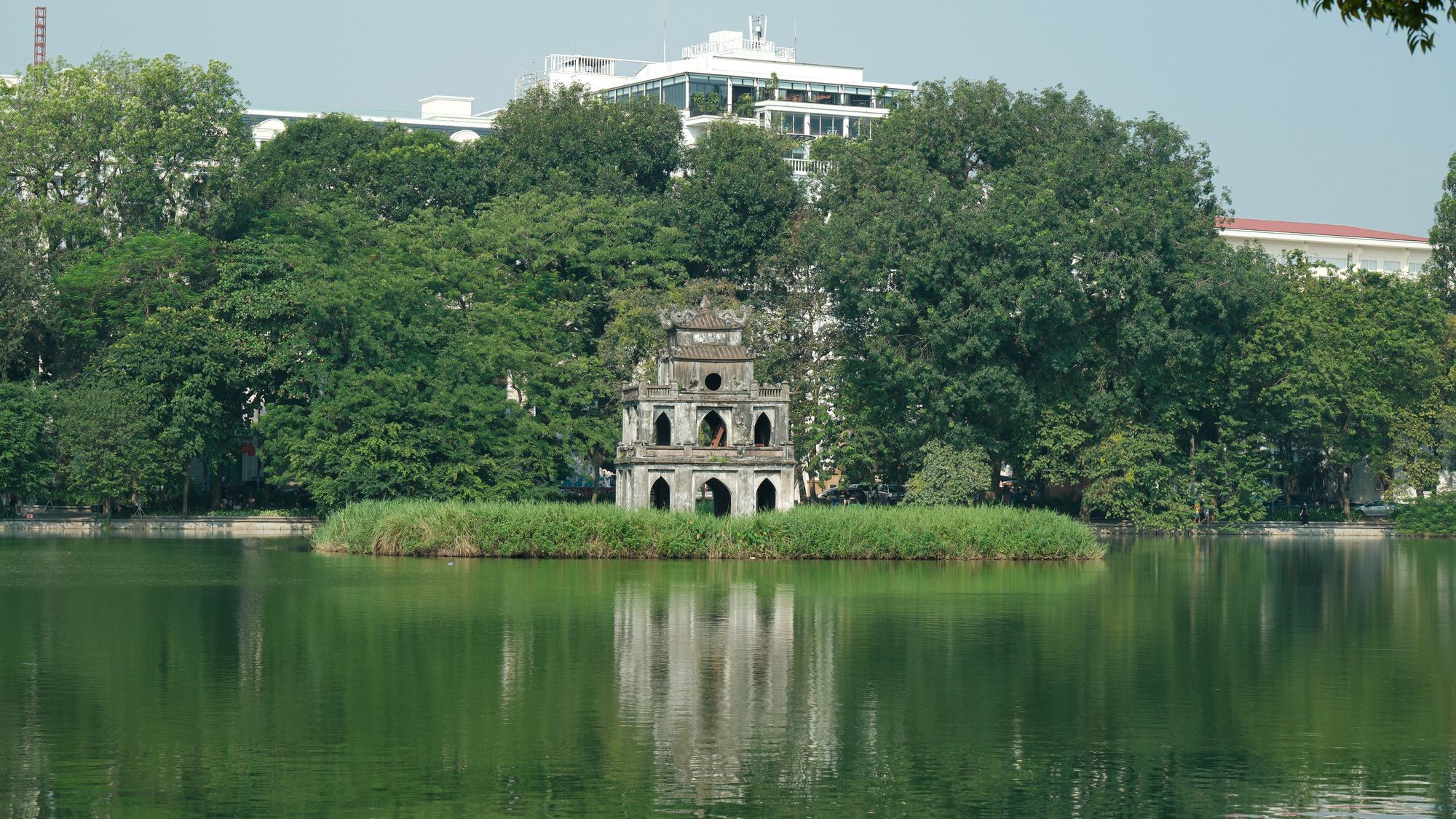 The Turtle Tower on an islet in Hoan Kiem Lake in Hanoi, surrounded by green trees, with the lake in the foreground.