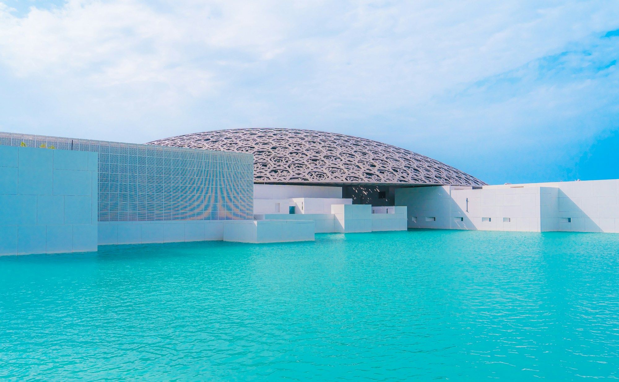 The lattice dome of the Louvre Abu Dhabi above the turquoise water that surrounds the museum, under a blue sky.