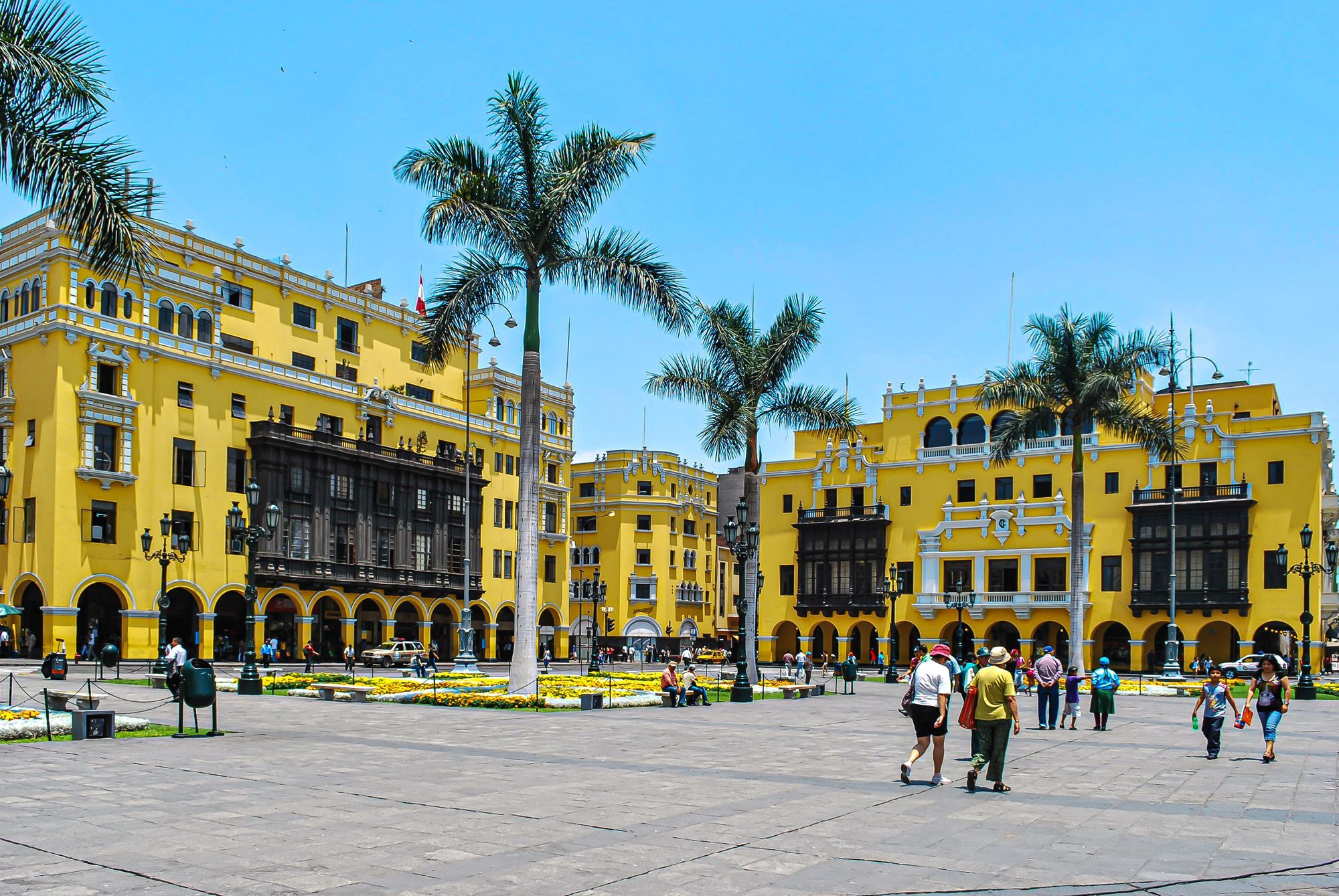 People walking across the Plaza Mayor in Lima, with palm trees and yellow colonial buildings.