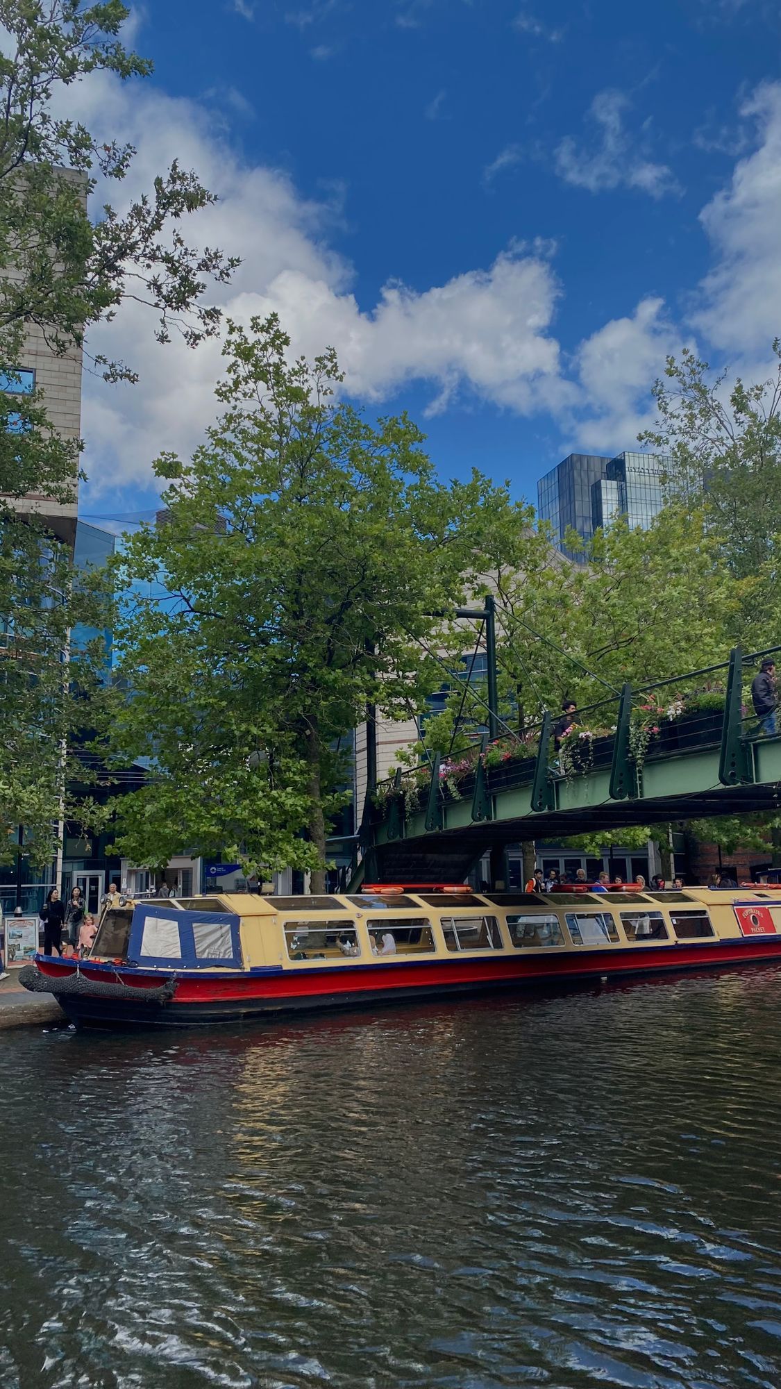 A narrowboat on a canal in Birmingham, with trees and an iron footbridge behind.
