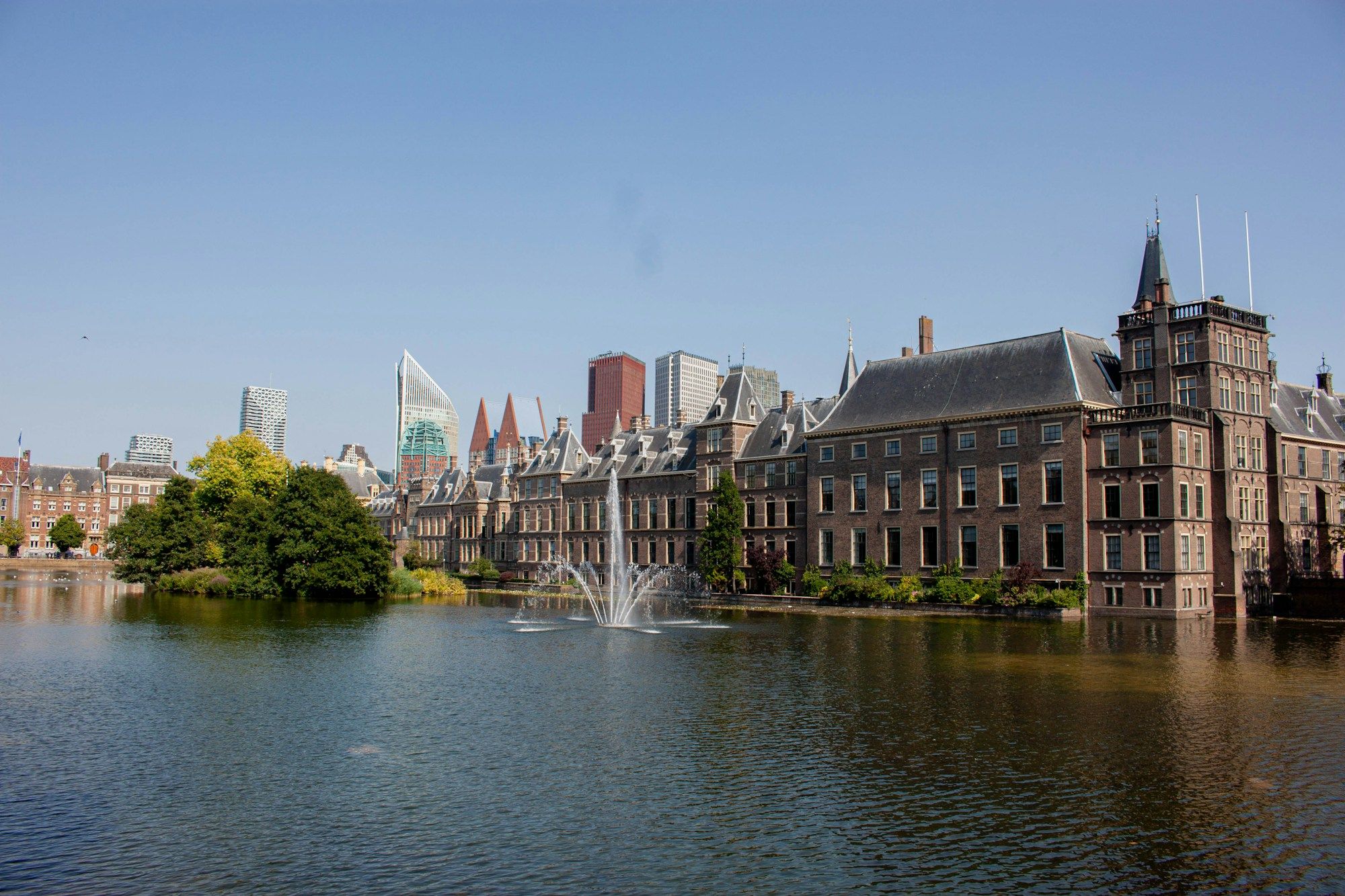 The Binnenhof buildings and the Hofvijver pond in The Hague, with a fountain and trees in front of the city skyline.