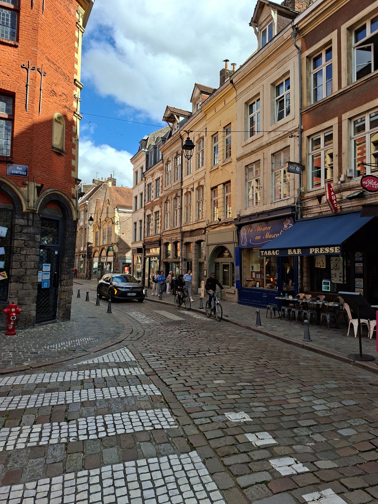 A cobbled street in the old town of Lille with a cafe terrace, a tabac and people on bicycles.