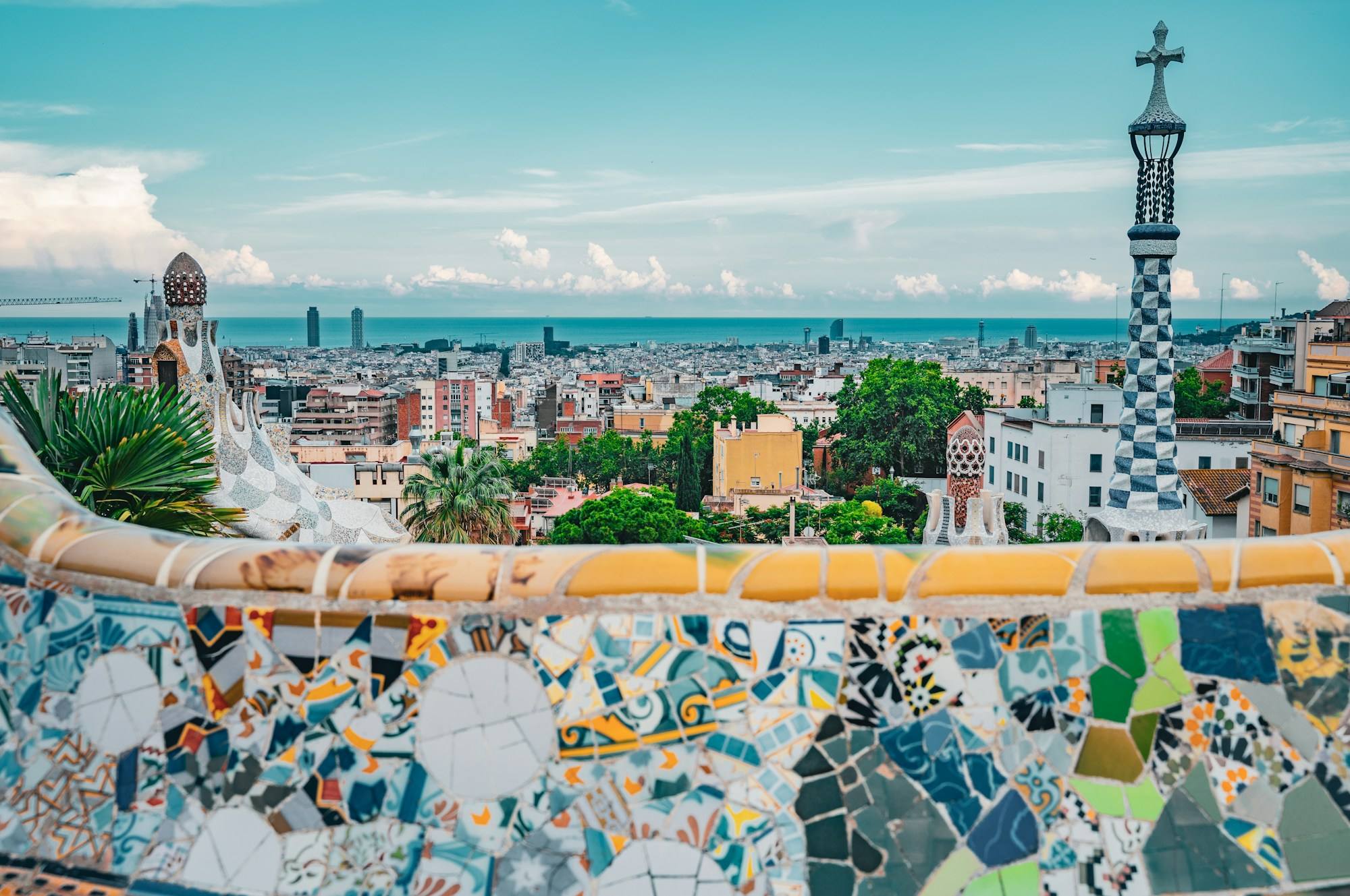 A mosaic tower in Park Güell, Barcelona, with the city and the Mediterranean behind it and a tiled bench in the foreground.