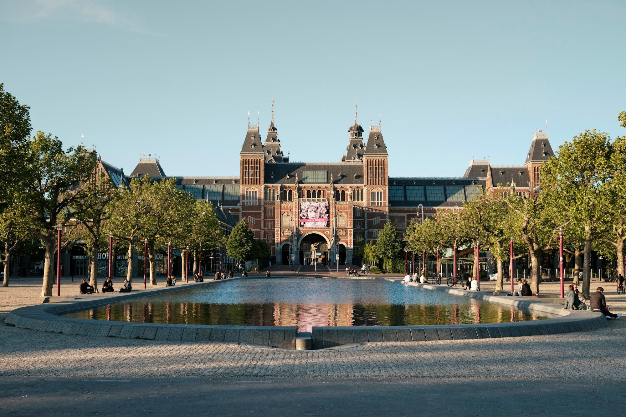 The Rijksmuseum in Amsterdam seen across its reflecting pool, with trees on both sides under a clear sky.