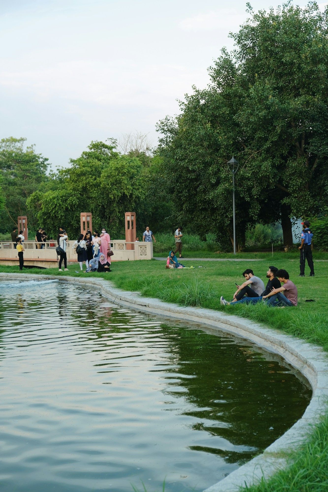 People sitting on the grass and walking beside a pond in Sunder Nursery park in New Delhi, with trees and a stone pavilion behind them.