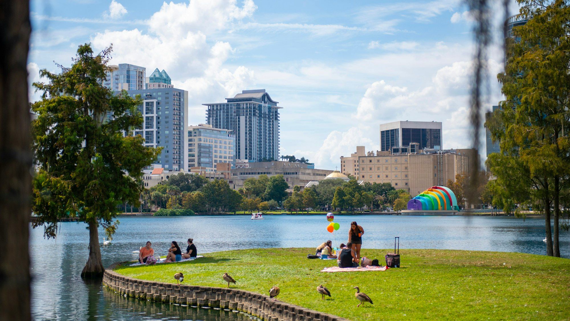 People sitting on the grass beside Lake Eola in Orlando, with downtown towers behind them across the water.