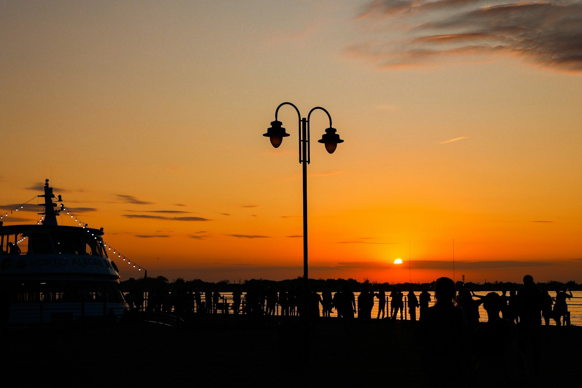 Pessoas em silhueta na grade da orla do Guaíba, em Porto Alegre, ao pôr do sol, ao lado de um poste de luz e de um barco com luzinhas.
