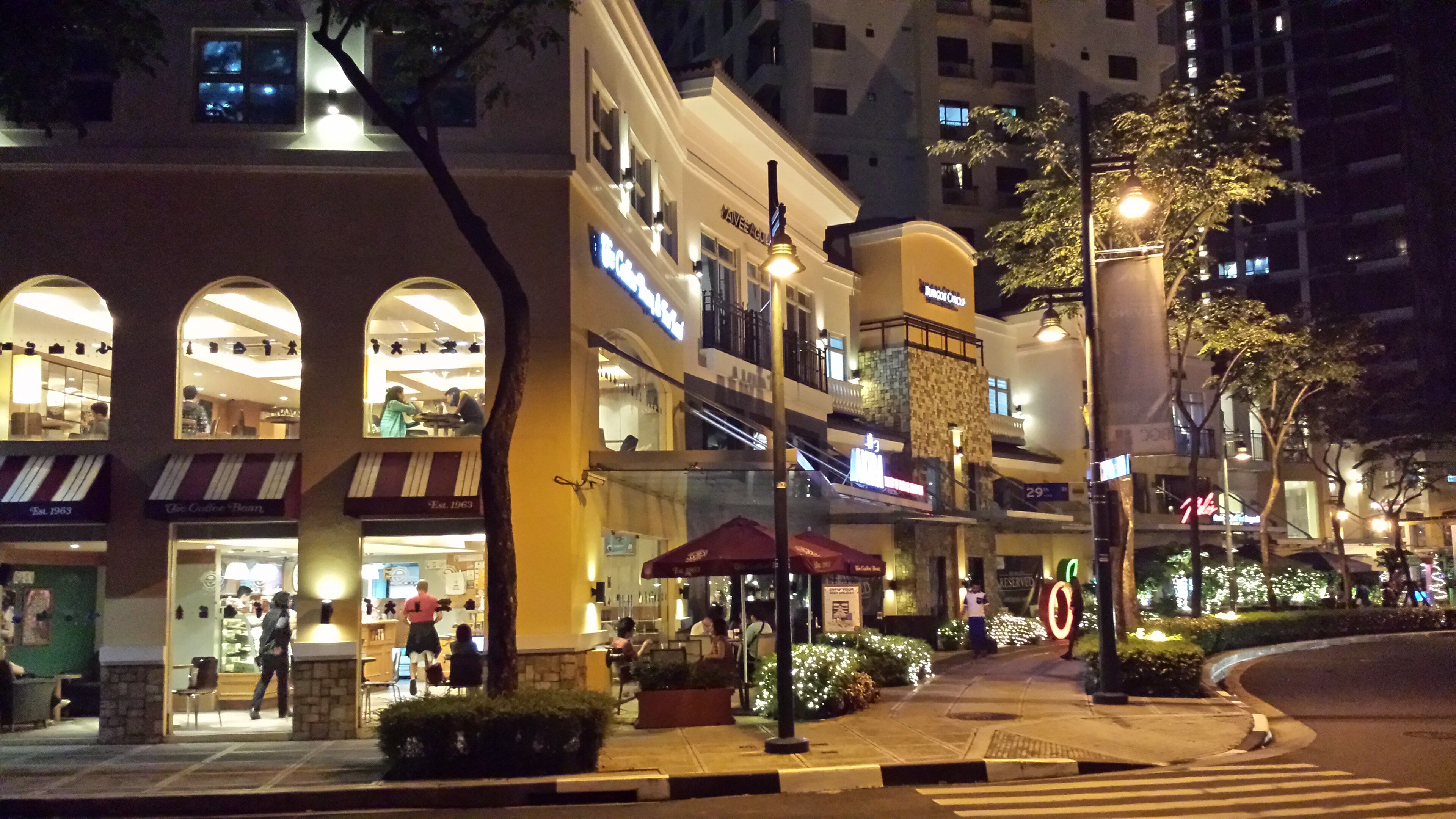 Cafes and restaurants lit up around Burgos Circle in Bonifacio Global City, with people sitting inside and walking along the sidewalk.