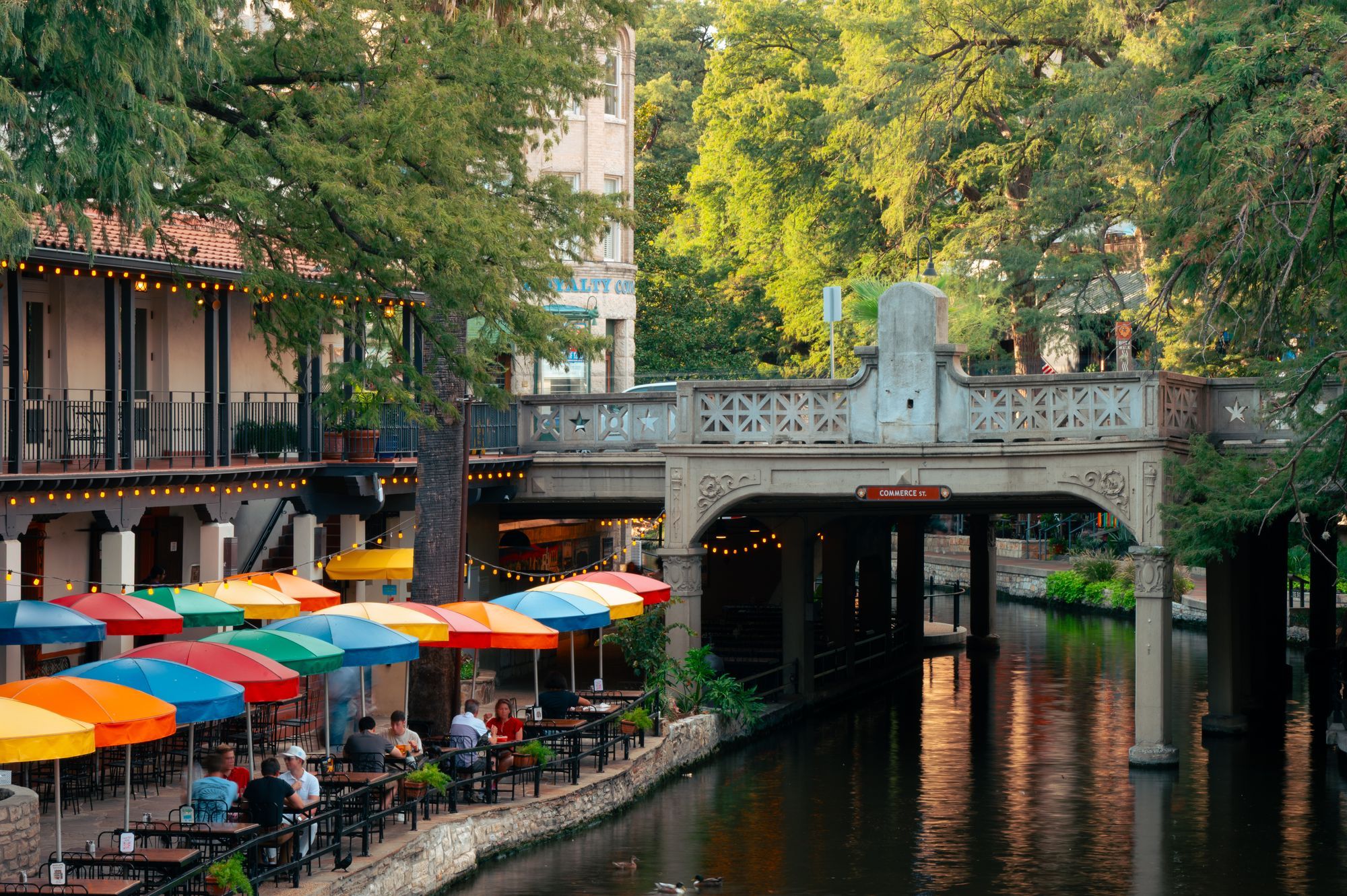 A stone bridge and outdoor tables with umbrellas beside the San Antonio River Walk.