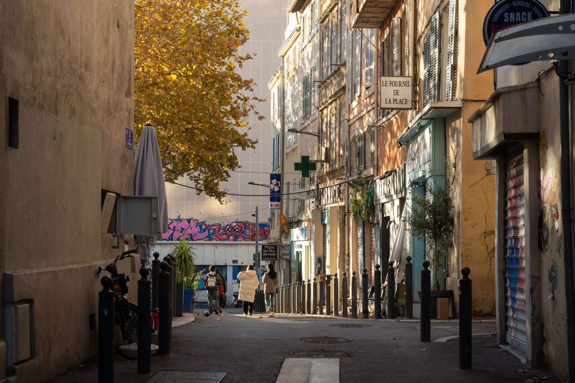 A narrow street in Marseille with people walking in warm afternoon light, a pharmacy cross and an autumn tree.
