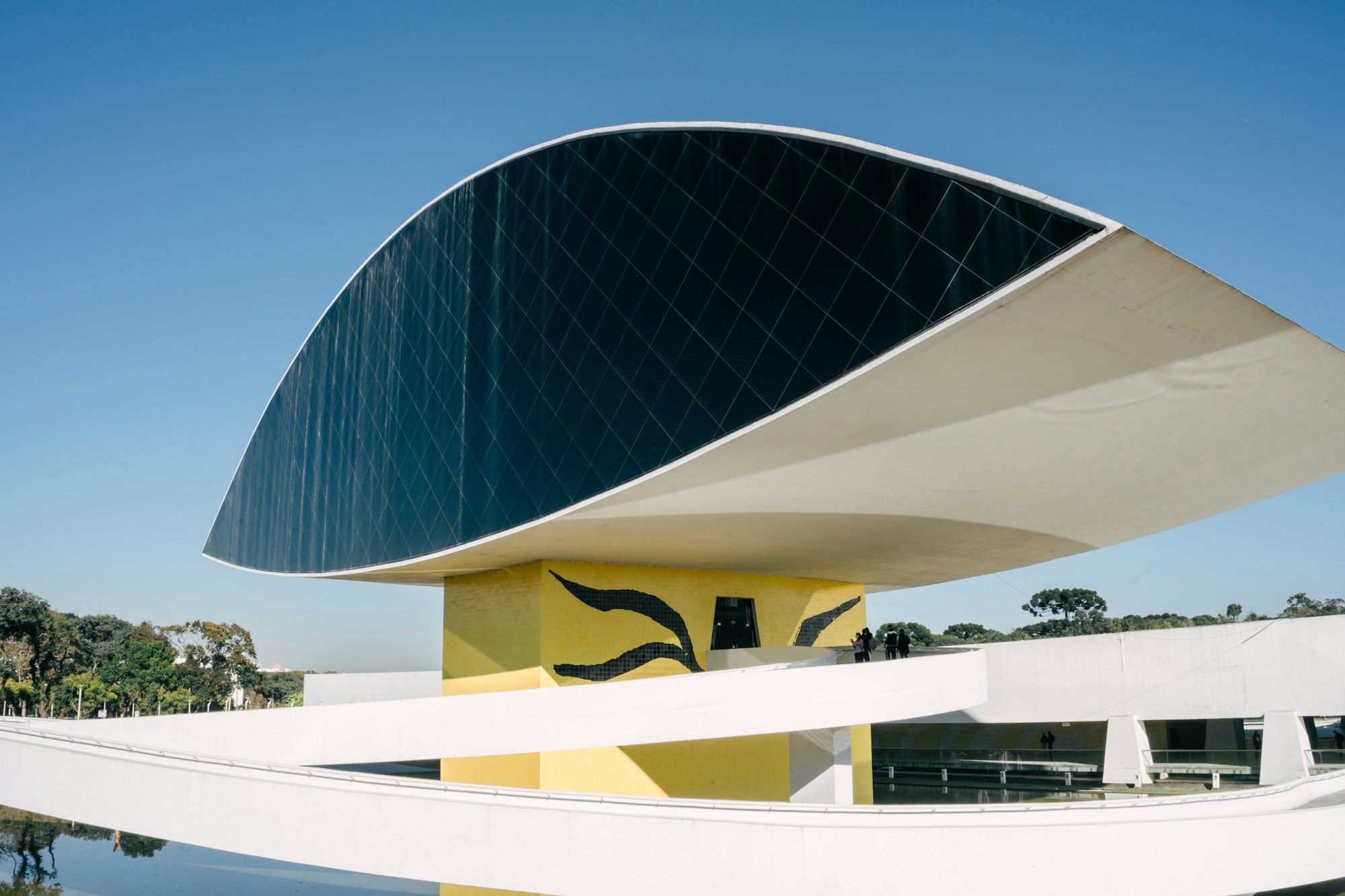 The Museu Oscar Niemeyer in Curitiba, with its eye-shaped glass volume above a yellow pillar, under a clear blue sky.
