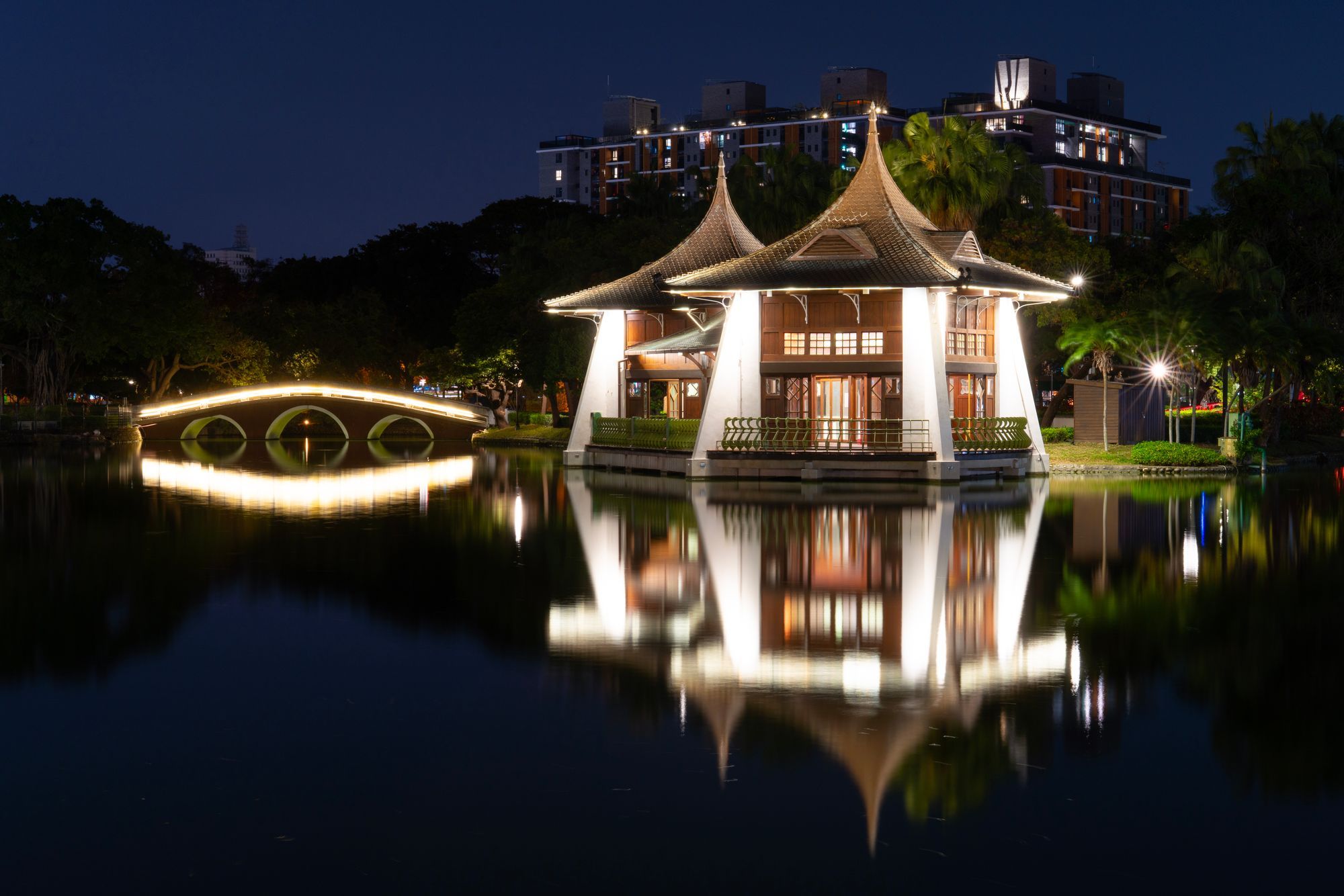 A lit lake pavilion and arched bridge reflected in a pond in Taichung.