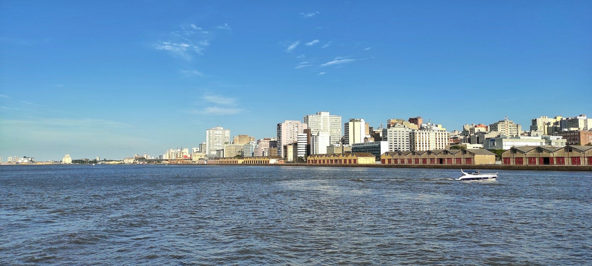 The Porto Alegre skyline and a row of low brick warehouses along the Guaíba, seen from the water under a blue sky, with a motorboat in the foreground.