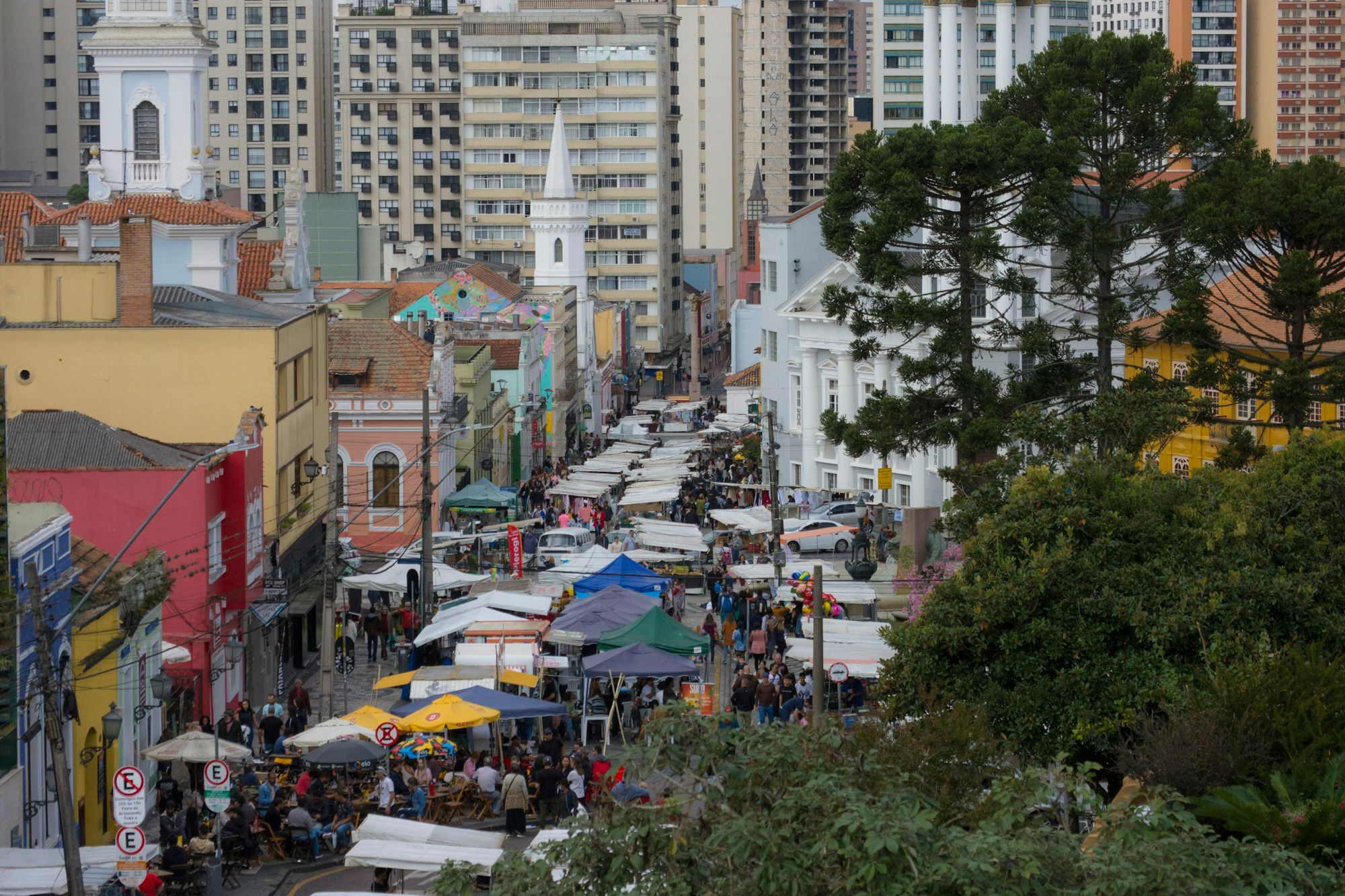 A street fair with stalls and crowded tables under umbrellas in central Curitiba, seen from above, with church towers and Araucaria trees on the right.
