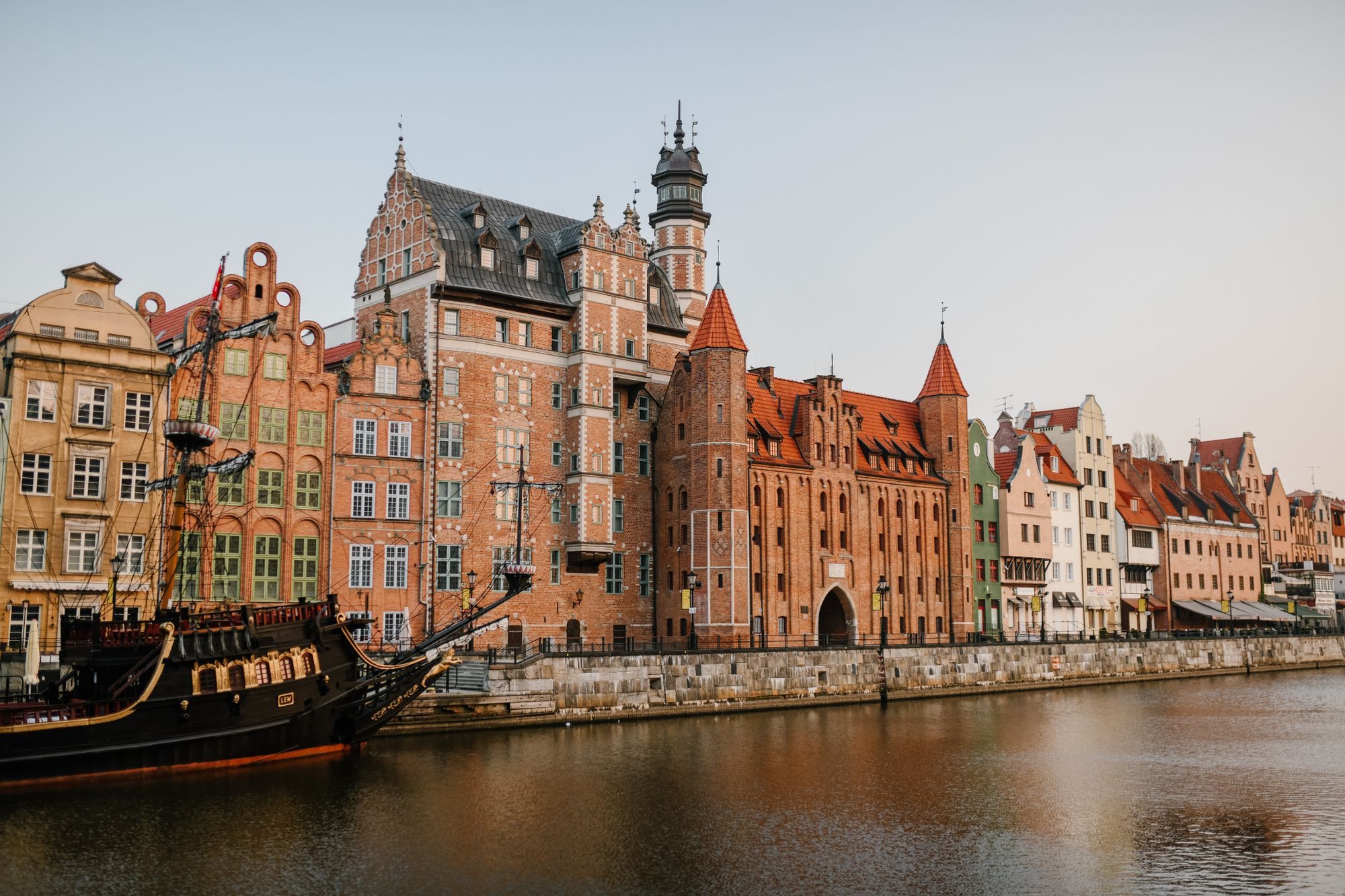Historic brick buildings along the Motława riverbank in Gdańsk, with a wooden tall ship moored at the left.