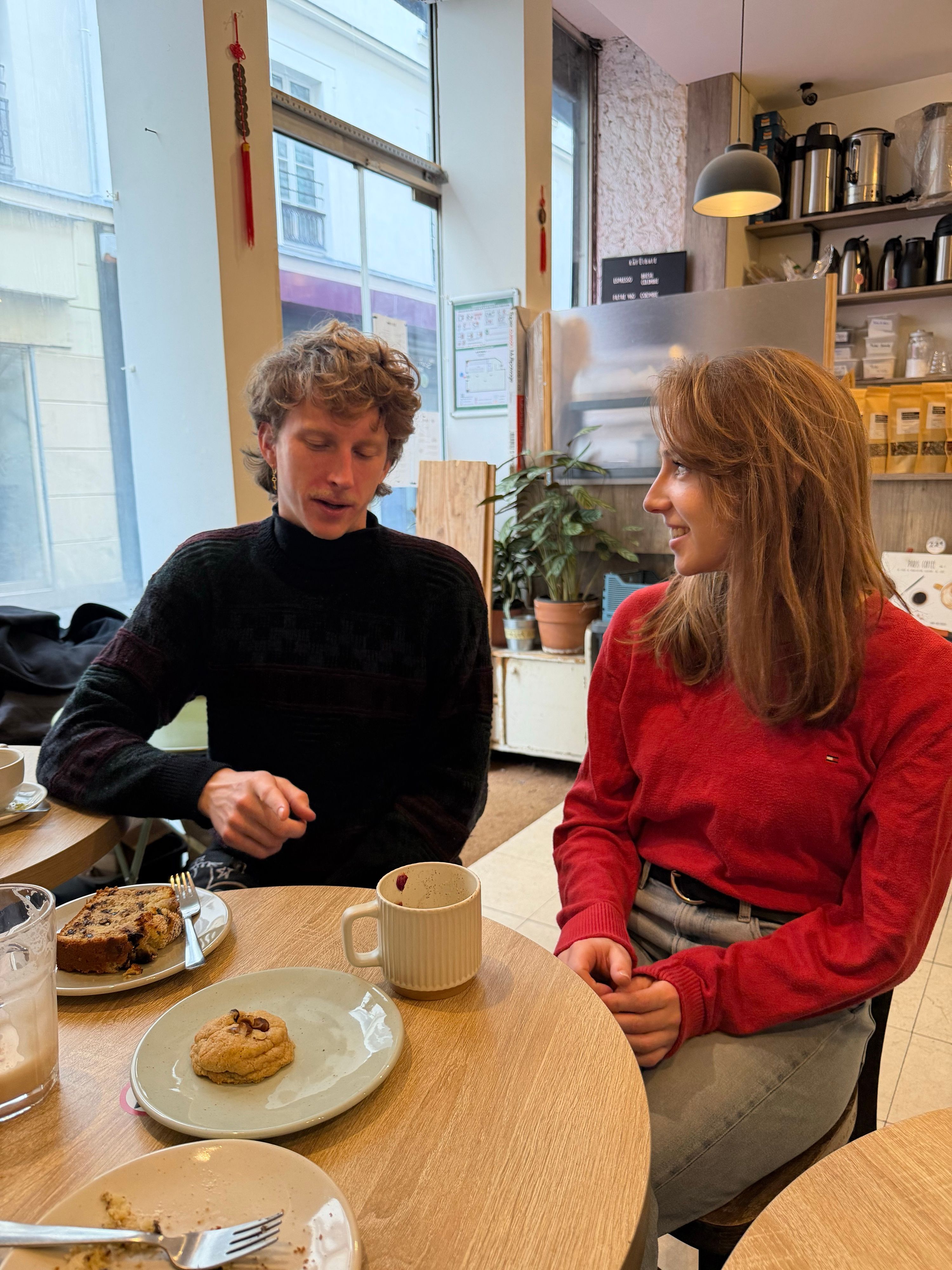 Two people talking over coffee and pastries at a table by a café window