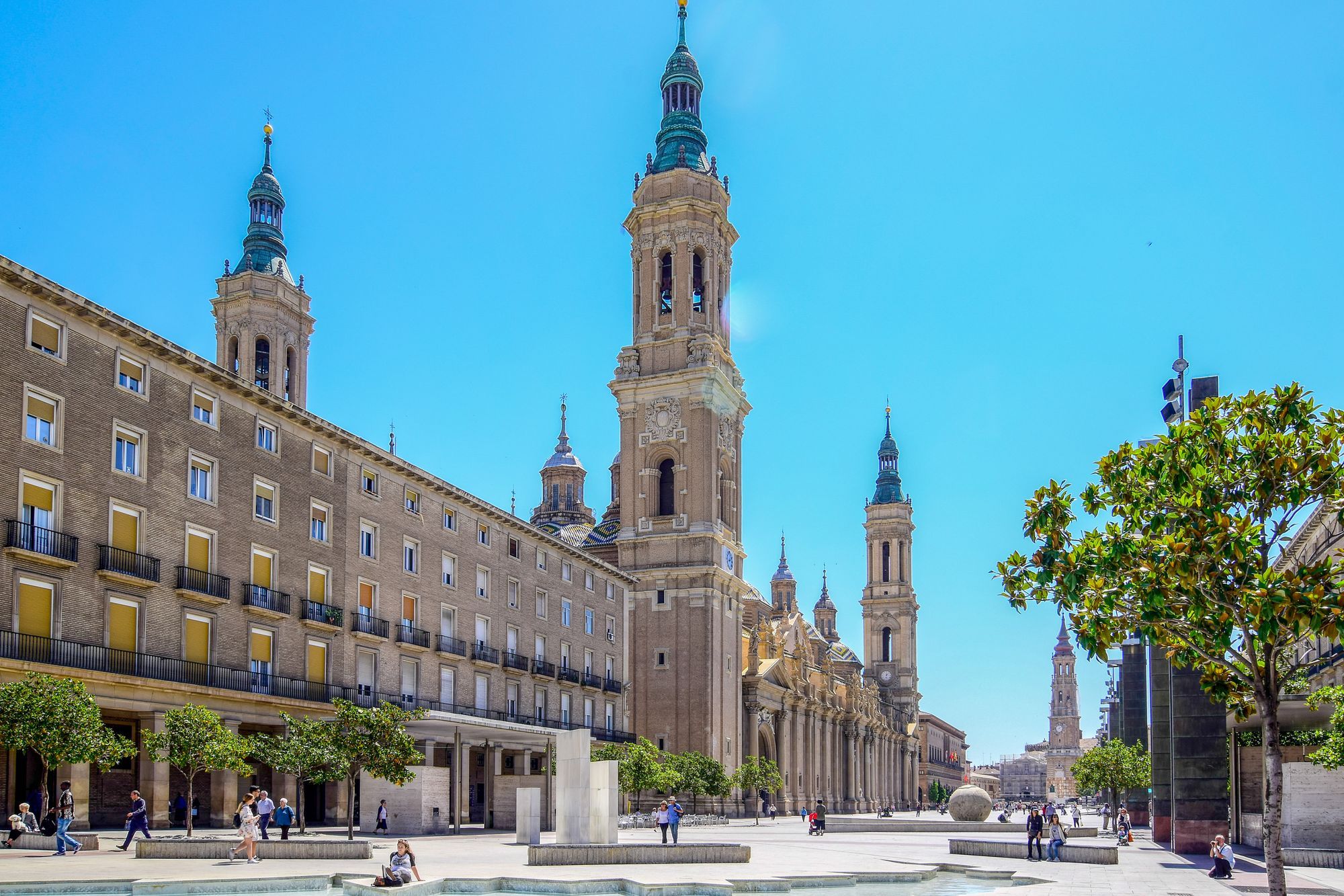 People crossing the Plaza del Pilar in Zaragoza, with the basilica towers and arcaded buildings beside it.