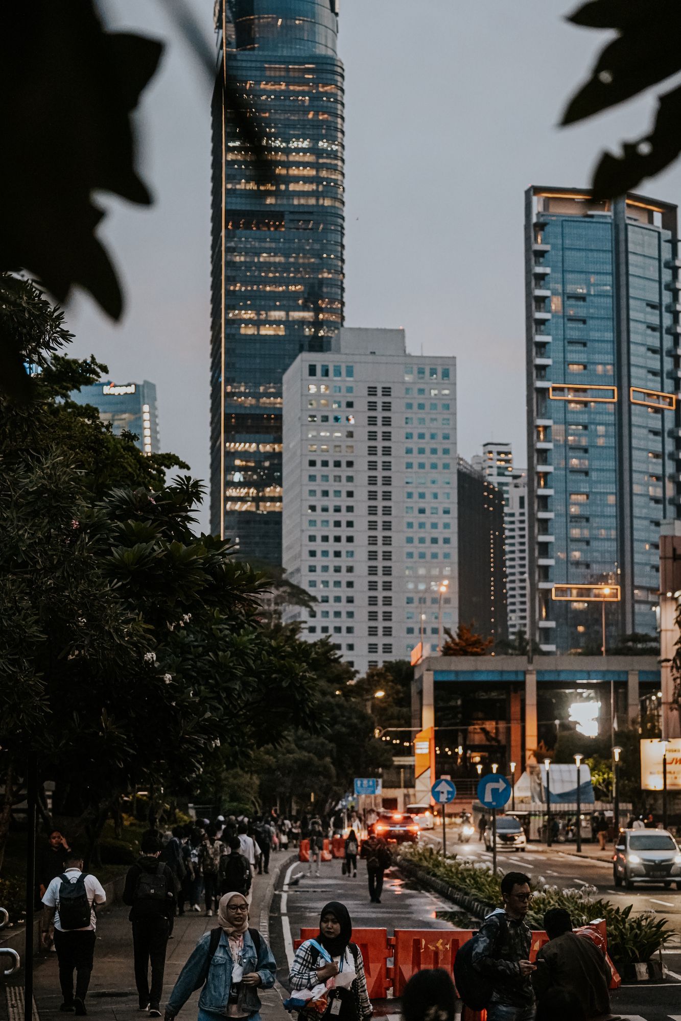 A busy street in Jakarta at dusk with pedestrians below office towers and trees.