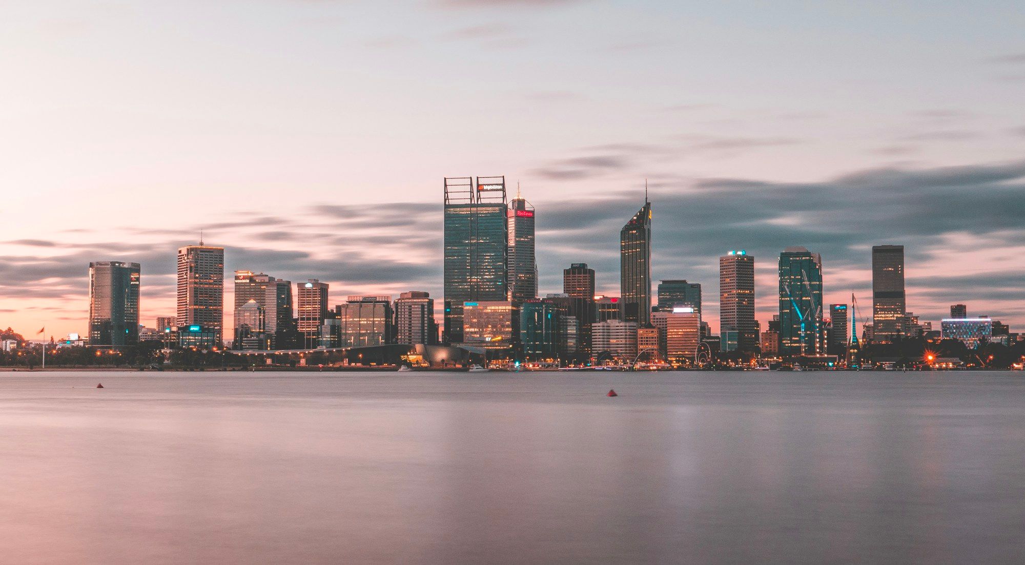 The Perth skyline across the Swan River at dusk, with calm water in the foreground.