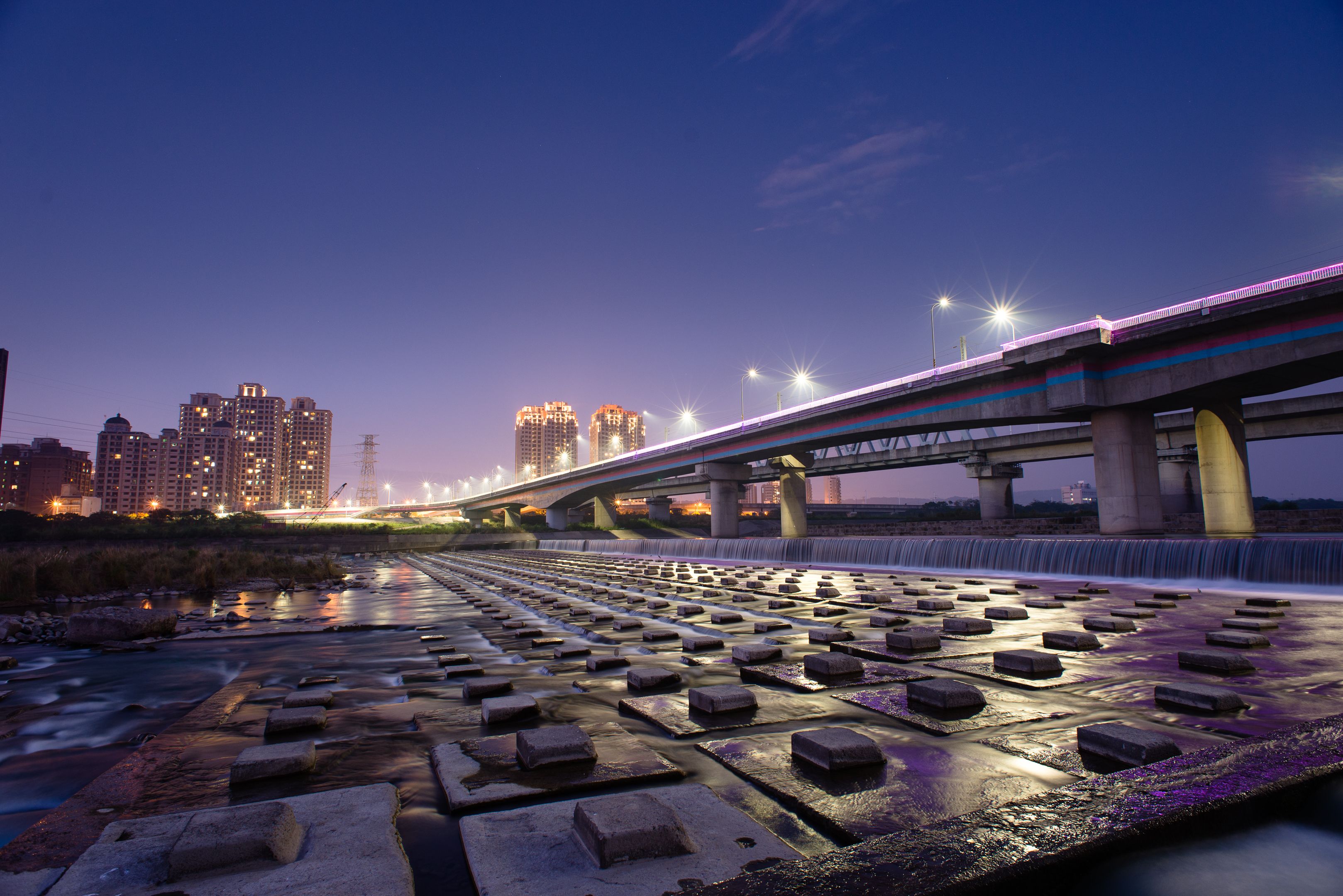 Apartment towers and a lit road bridge over a river weir in Hsinchu at dusk.