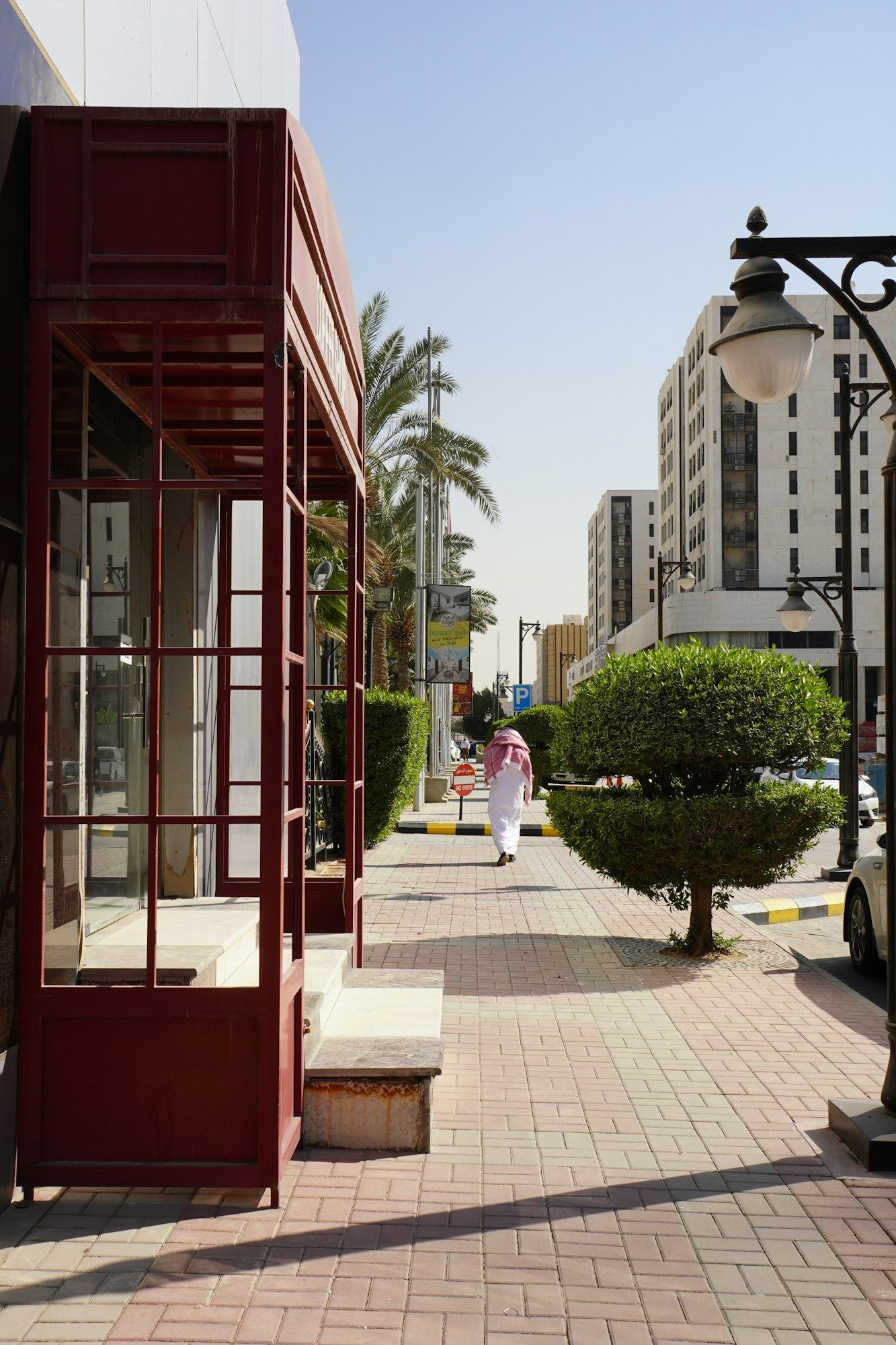 A sunlit sidewalk in Riyadh with a clipped bush, palm trees and a man in a white thobe walking away down the street.