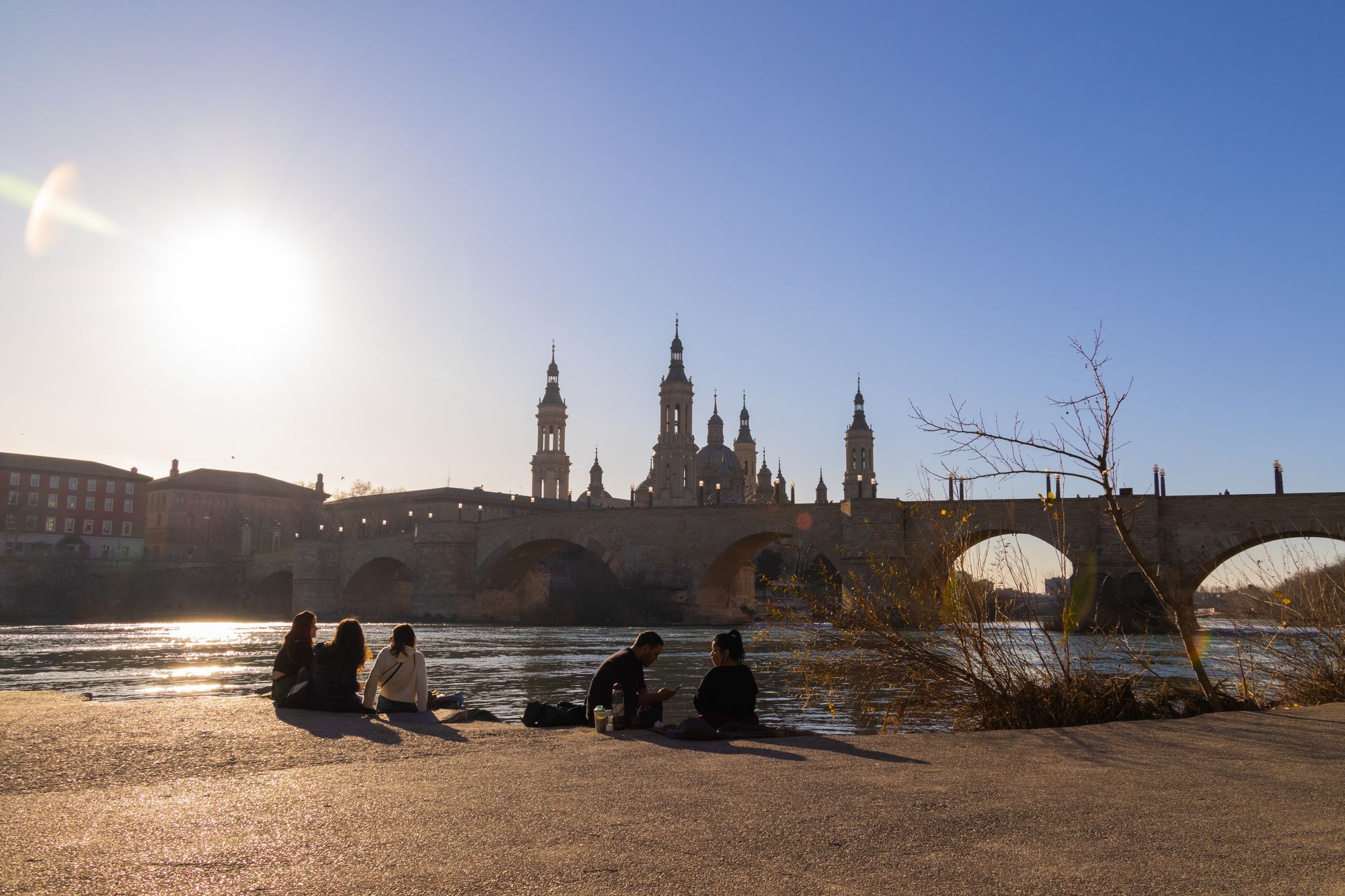 People sitting by the Ebro in Zaragoza, with the Stone Bridge and the basilica towers against the low sun.
