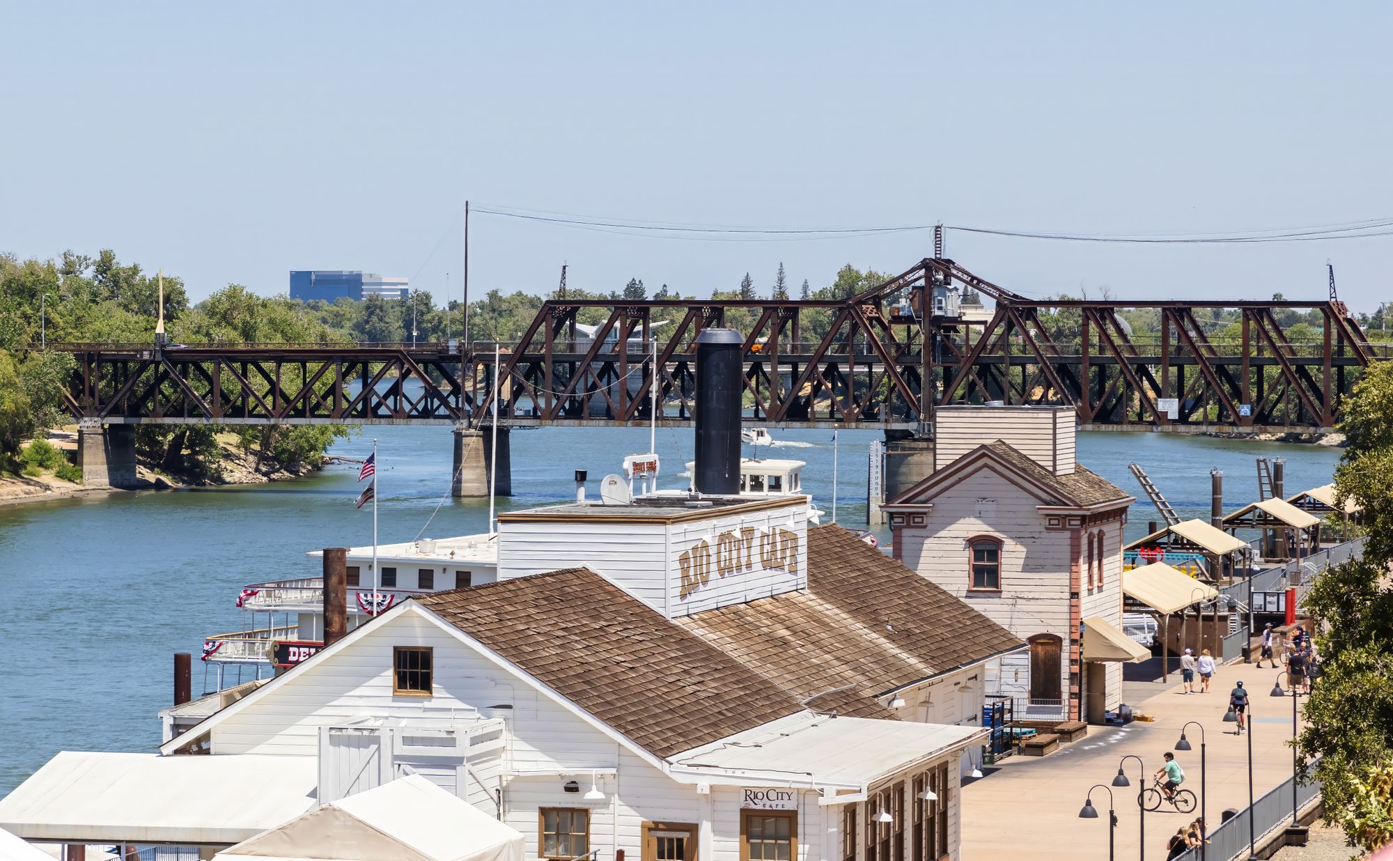 Old Sacramento's waterfront buildings and boat landing beside the river, with a rail bridge behind.