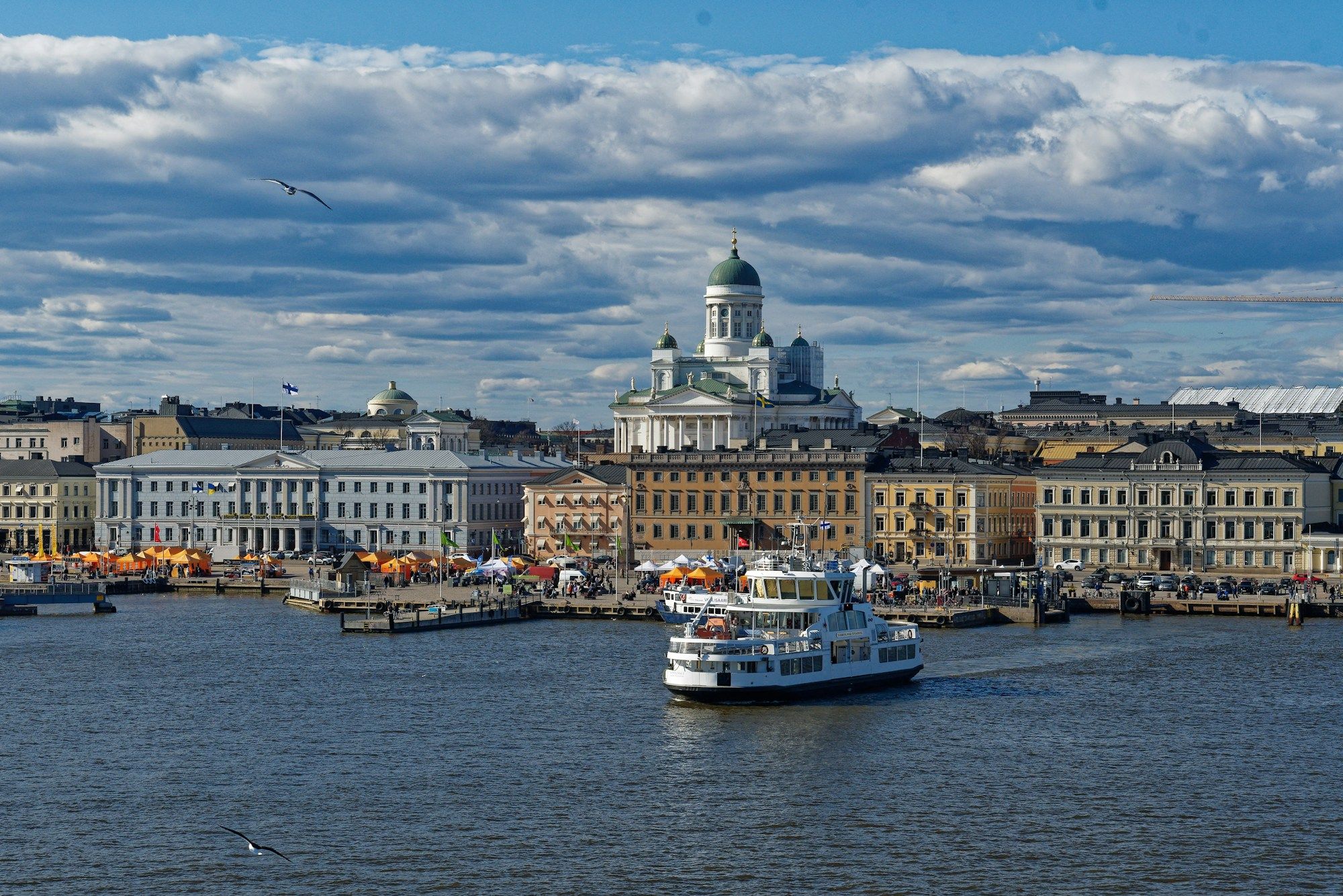 A white ferry on the water in front of the Helsinki waterfront, with Helsinki Cathedral rising above the buildings under a cloudy sky.